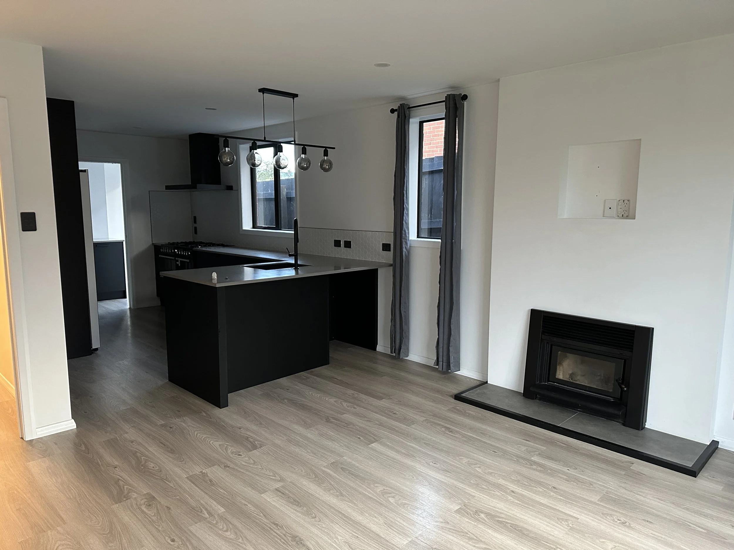 Empty living room with light wood flooring, white walls, and a black fireplace. There are two windows with gray curtains, a modern black and white kitchen island with a hanging light fixture above it, and black appliances, including a range hood.