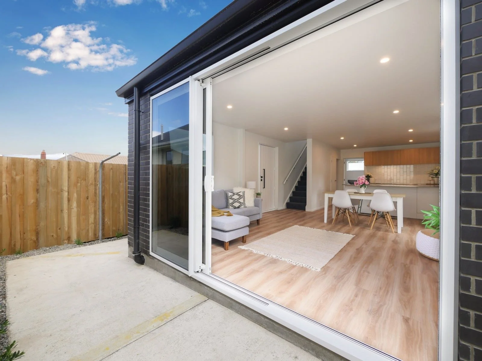 View of a modern, open-concept living and dining area with hardwood flooring, white walls, and kitchen in the background, seen through a large sliding glass door from an outdoor patio.