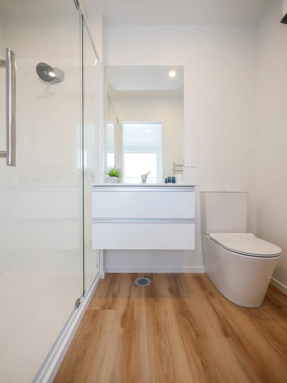 Modern bathroom with wooden floor, white vanity with mirror, plant, and toilet