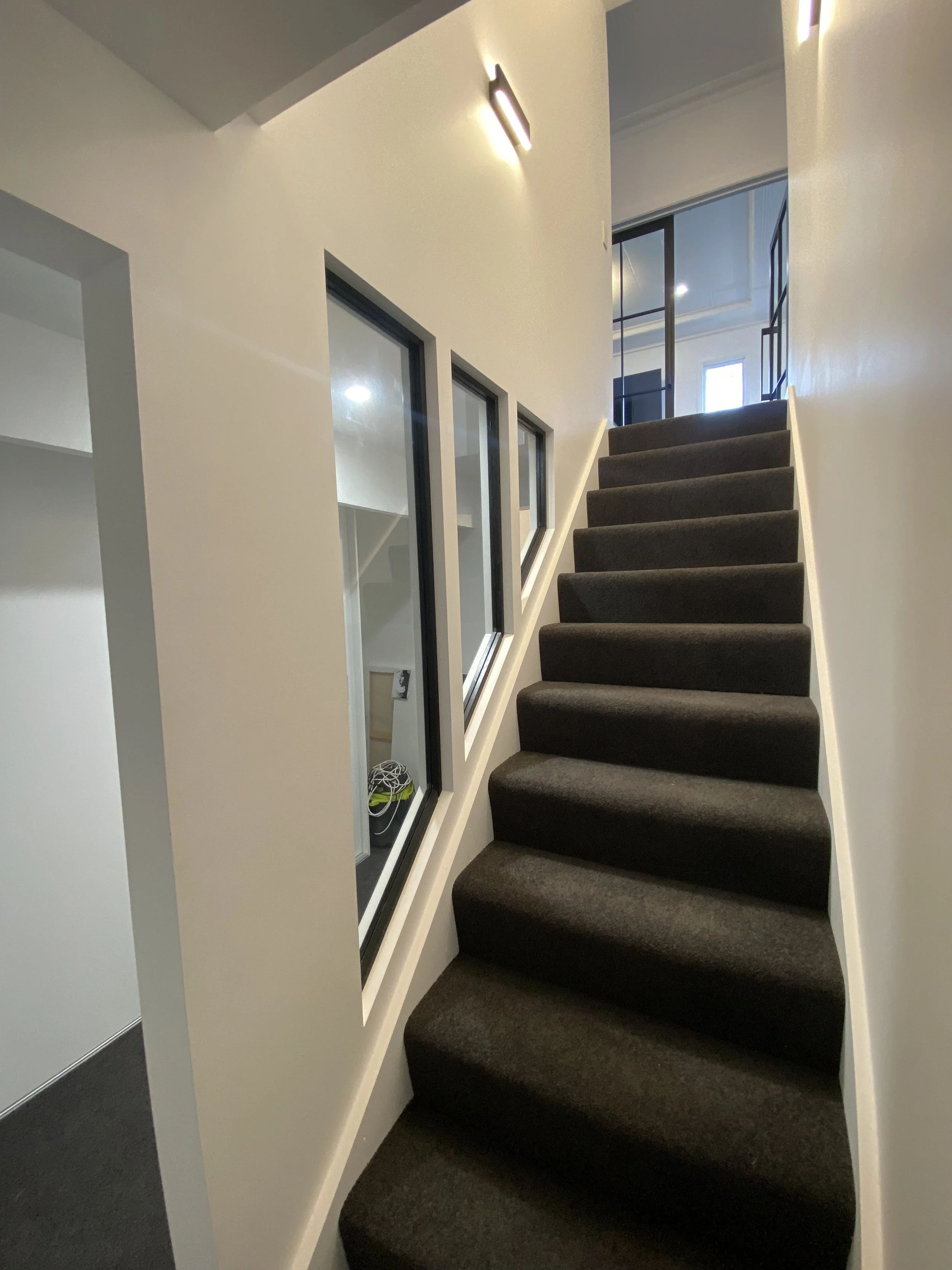 Indoor staircase with black carpeted steps, white walls, a wall-mounted light fixture, and windows with black frames at the top of the stairs.