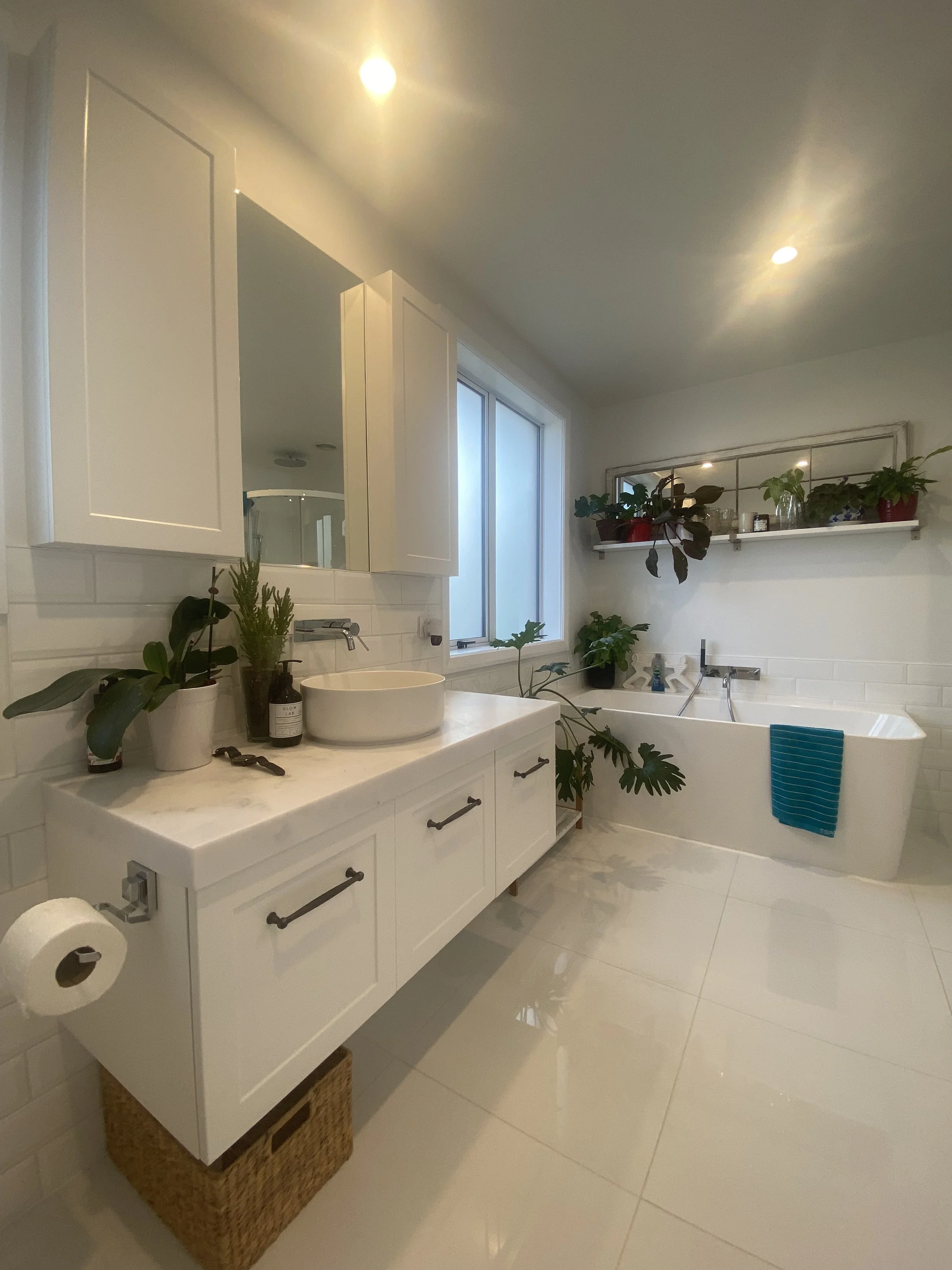 A modern bathroom with white cabinetry and a large mirror, decorated with green plants. There is a bathtub with a blue towel hanging on its edge, and a window providing natural light. The bathroom features white walls, tiles, and a clean, minimalist 