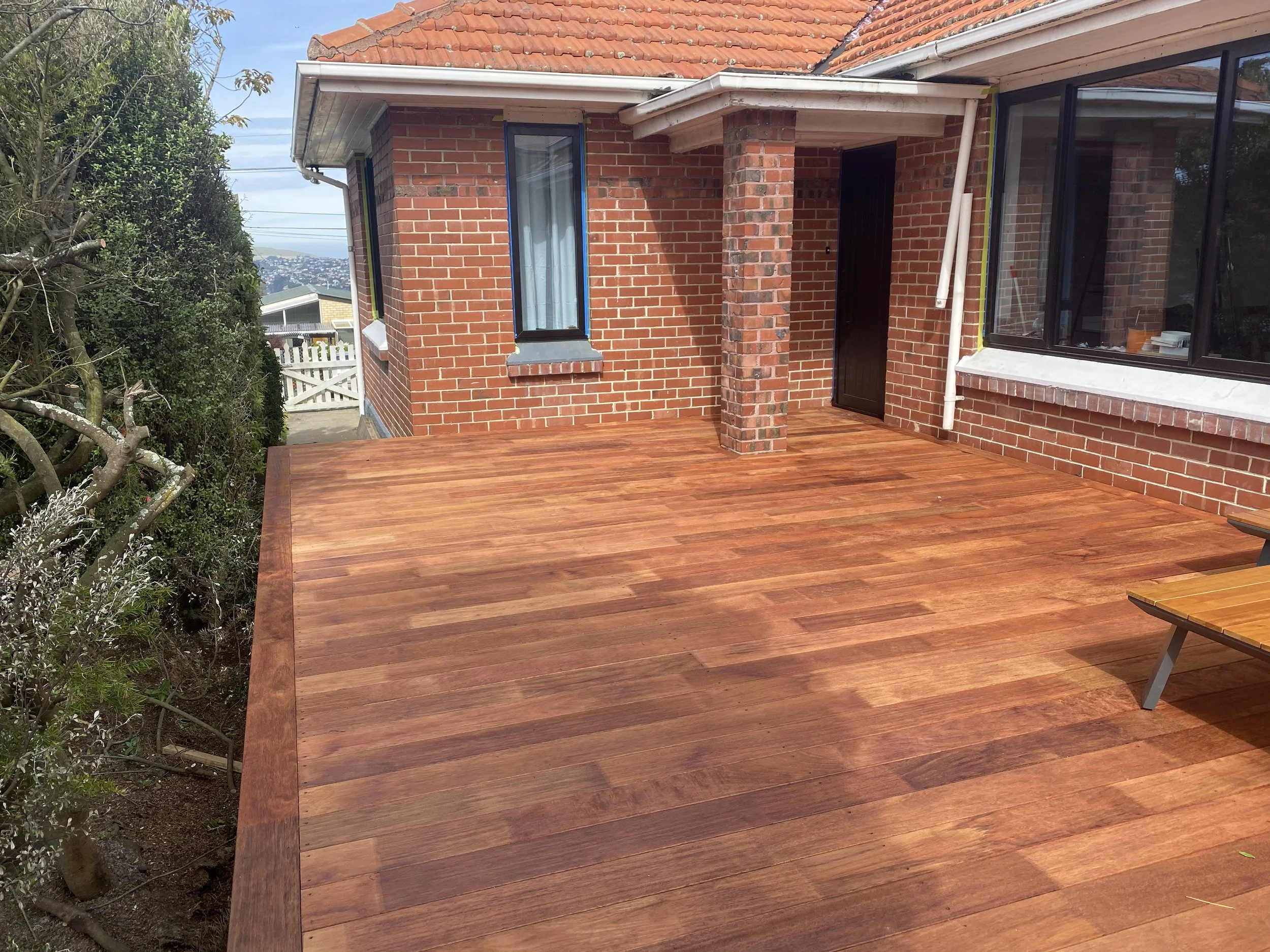 A backyard deck made of wooden planks attached to a brick house with large windows and a small brick column. The house has a sloped tile roof, and there is a tree and bushes on the left side of the image, with a view of a neighborhood and hills in th