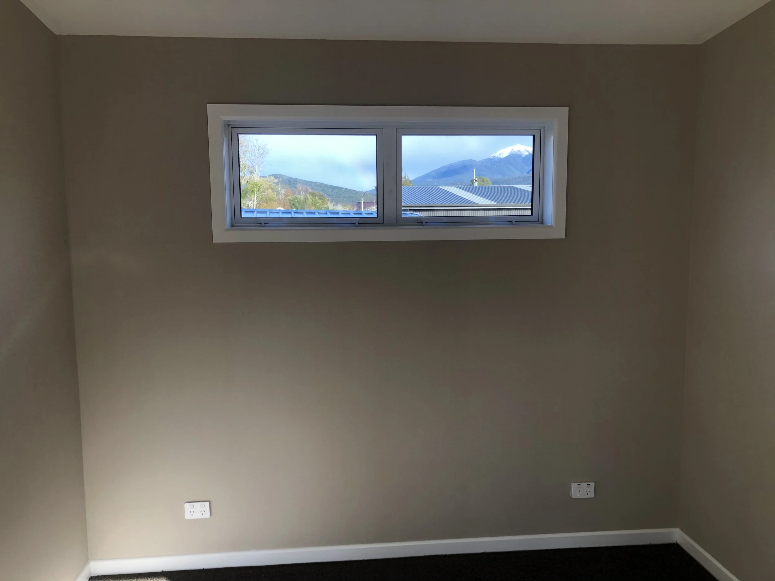 View of a window on a beige-colored wall showing mountains and blue sky outside, with two electrical outlets near the floor.