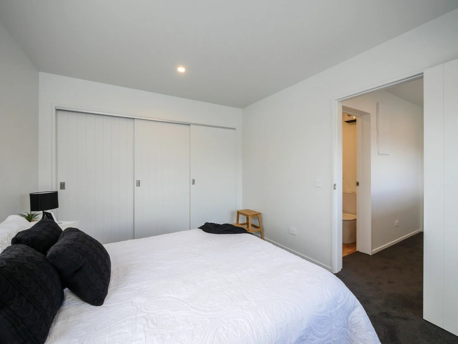 A bedroom with a white bedspread, black pillows, a black lamp, and some small plants on a white nightstand. The room has a closet with white sliding doors, a small wooden stool, and an open doorway leading to a bathroom.