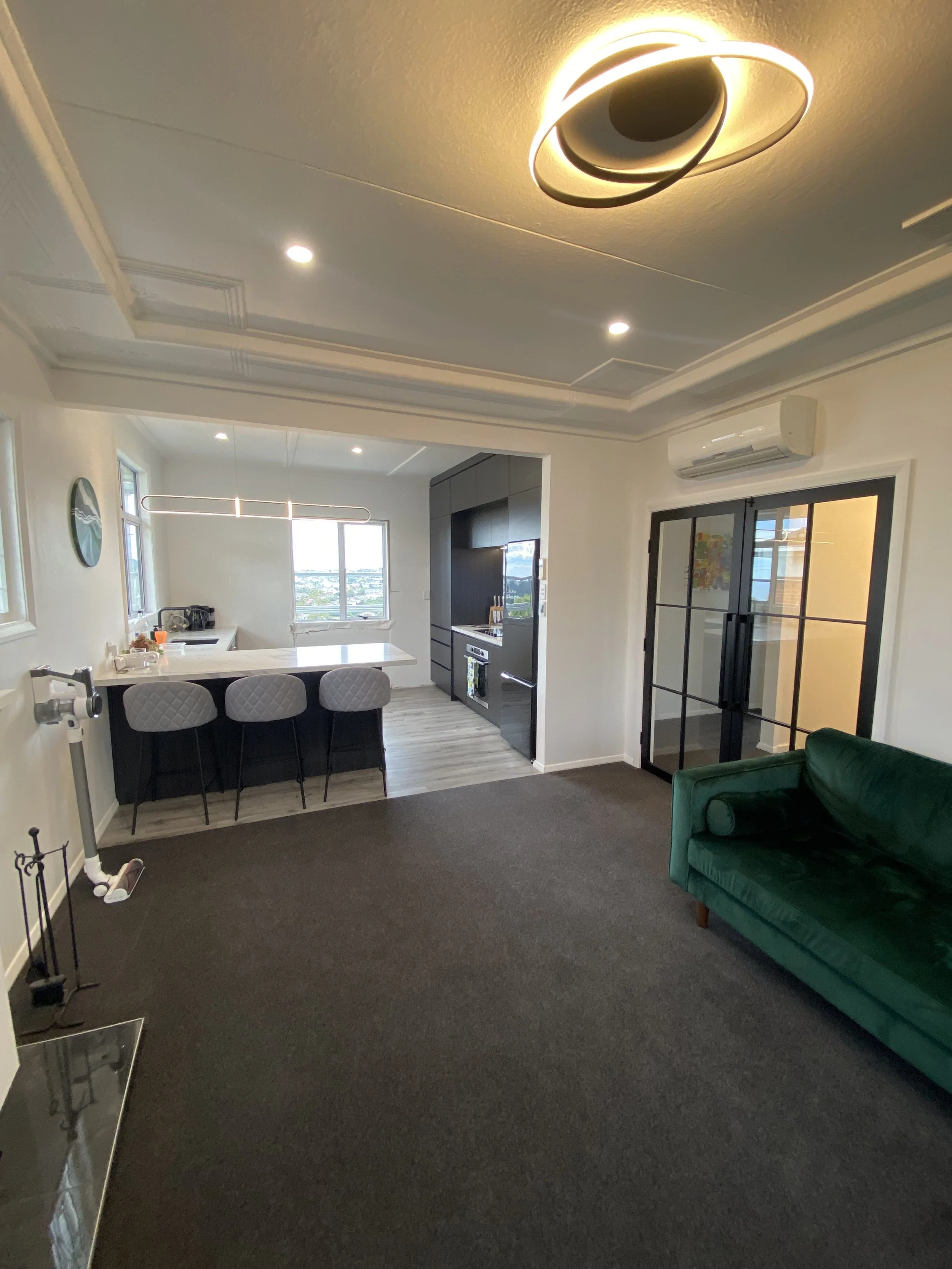 Interior of a modern living room with a green sofa, black glass door, a kitchen area in the background, and two contemporary light fixtures on the ceiling. There is an air conditioning unit above the door and a view outside through a window.