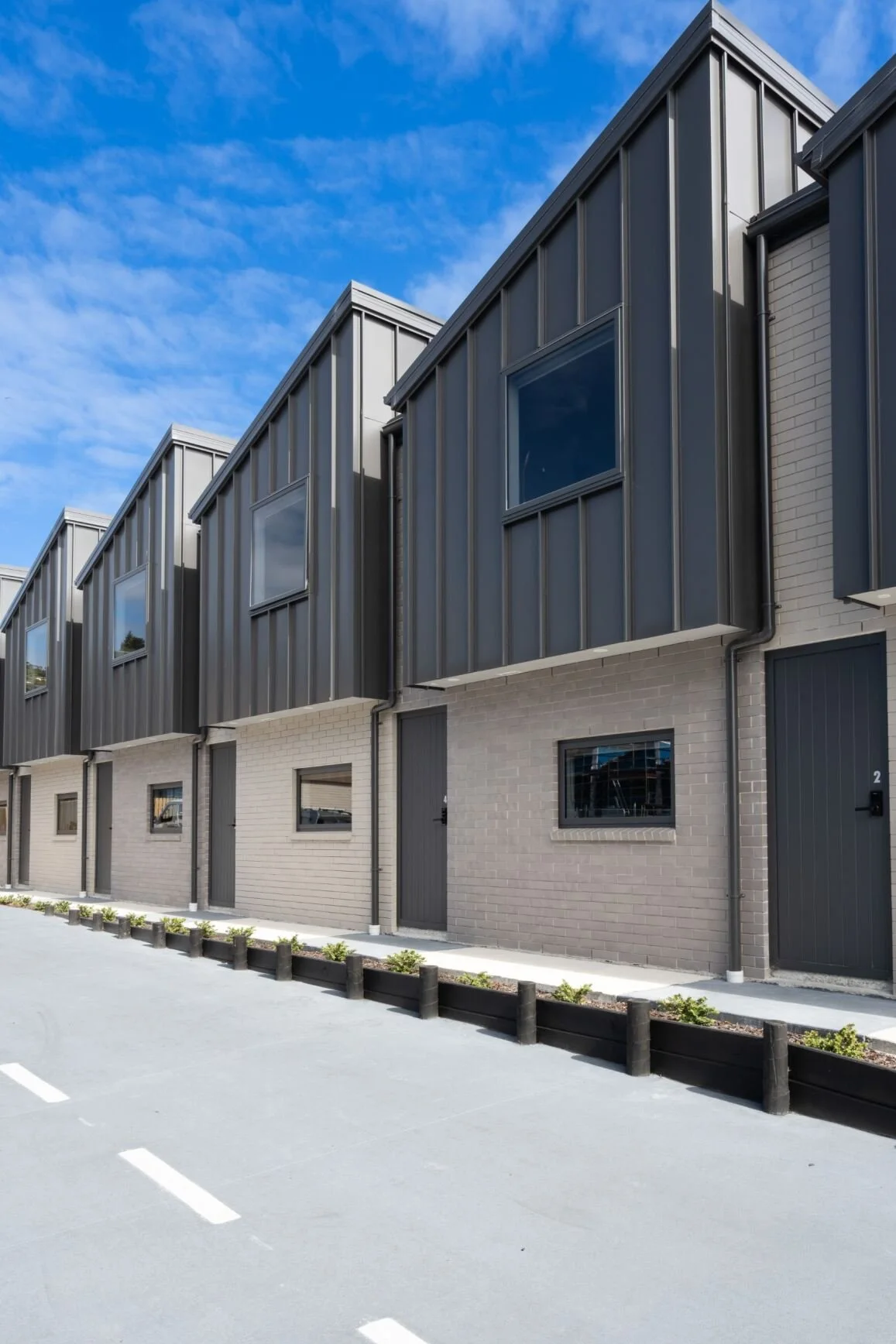 Modern townhouses with metal and brick facades, parking lot with marked spaces, and a partly cloudy sky.