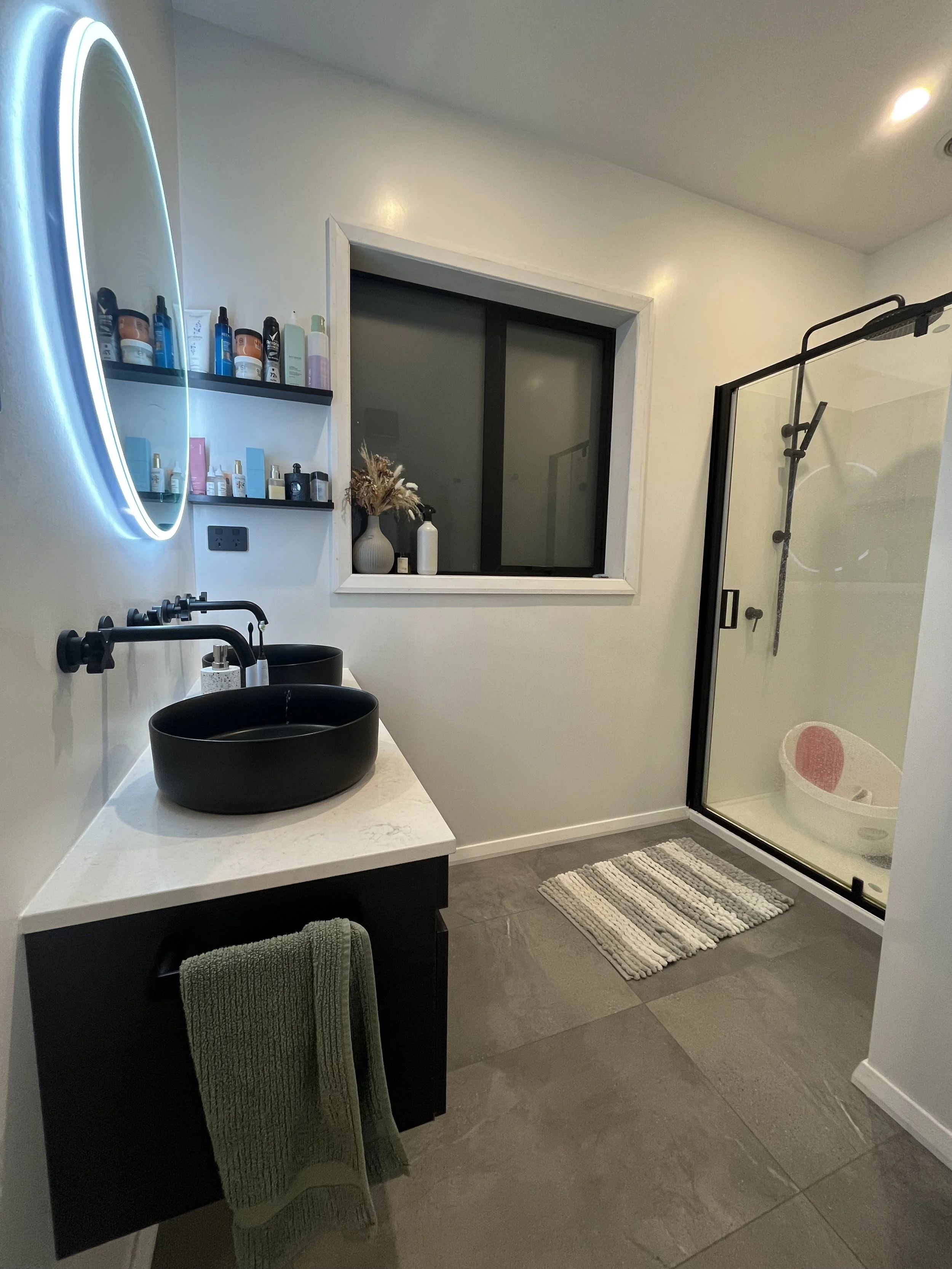 Modern bathroom with black vessel sinks, a large mirror with LED lighting, open shelves with toiletries, a window, a vase with dried flowers, a shower with black fixtures, and a striped bath mat.
