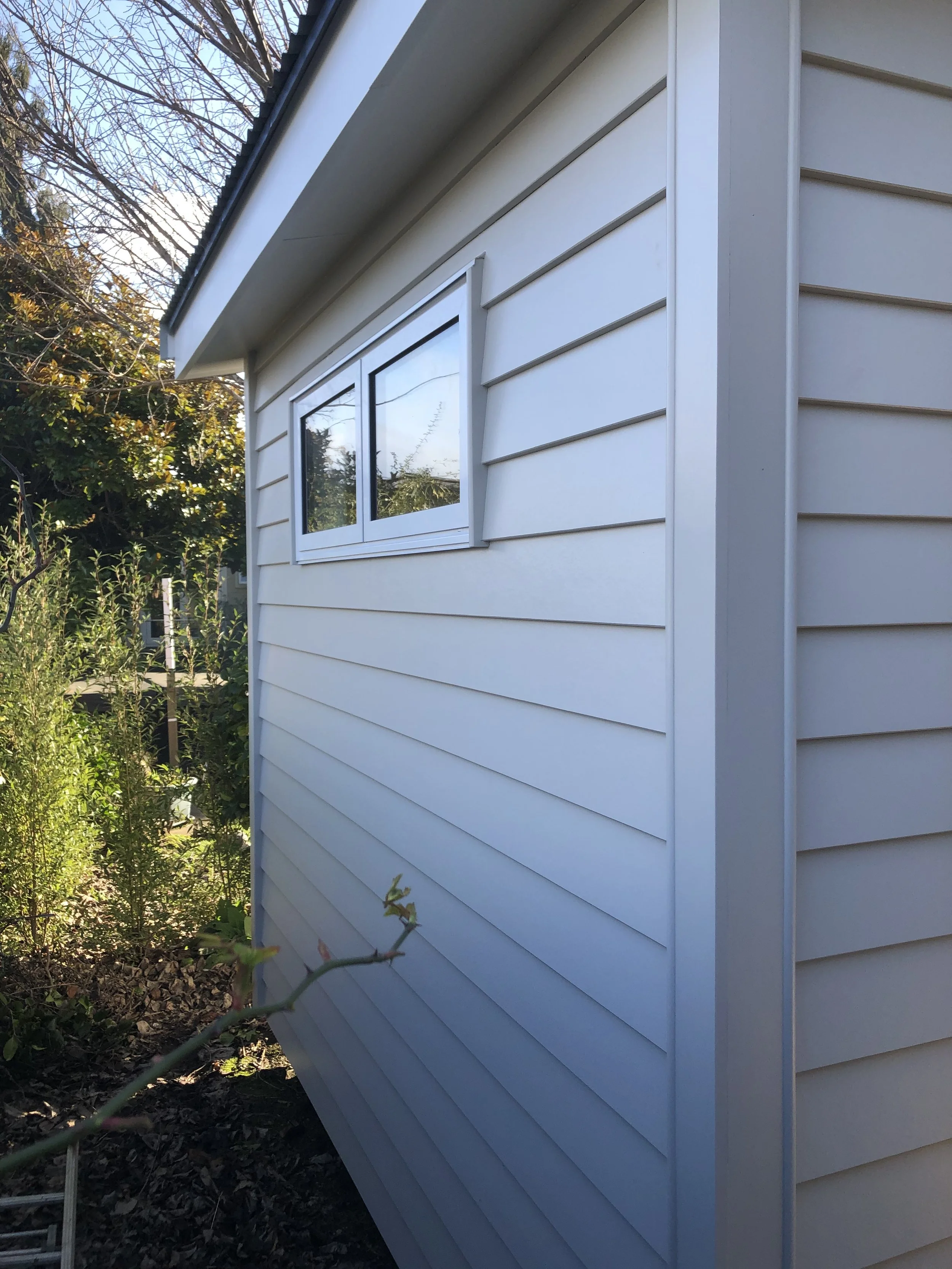 Side view of a white house wall with horizontal siding and a small rectangular window, surrounded by greenery and trees, under a partly cloudy sky.