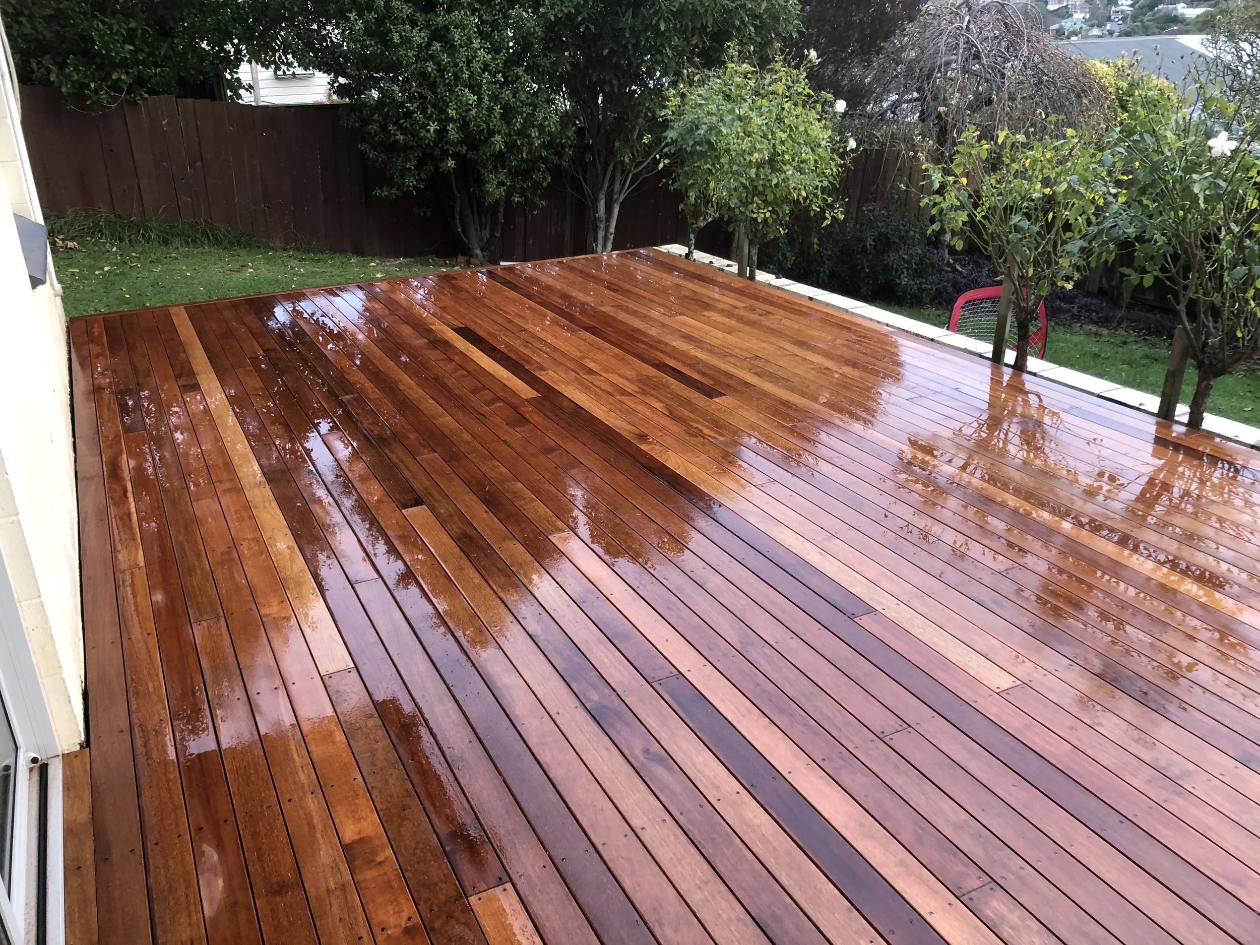A wet wooden deck outside a house with trees, a red fence, and a red chair in the background.