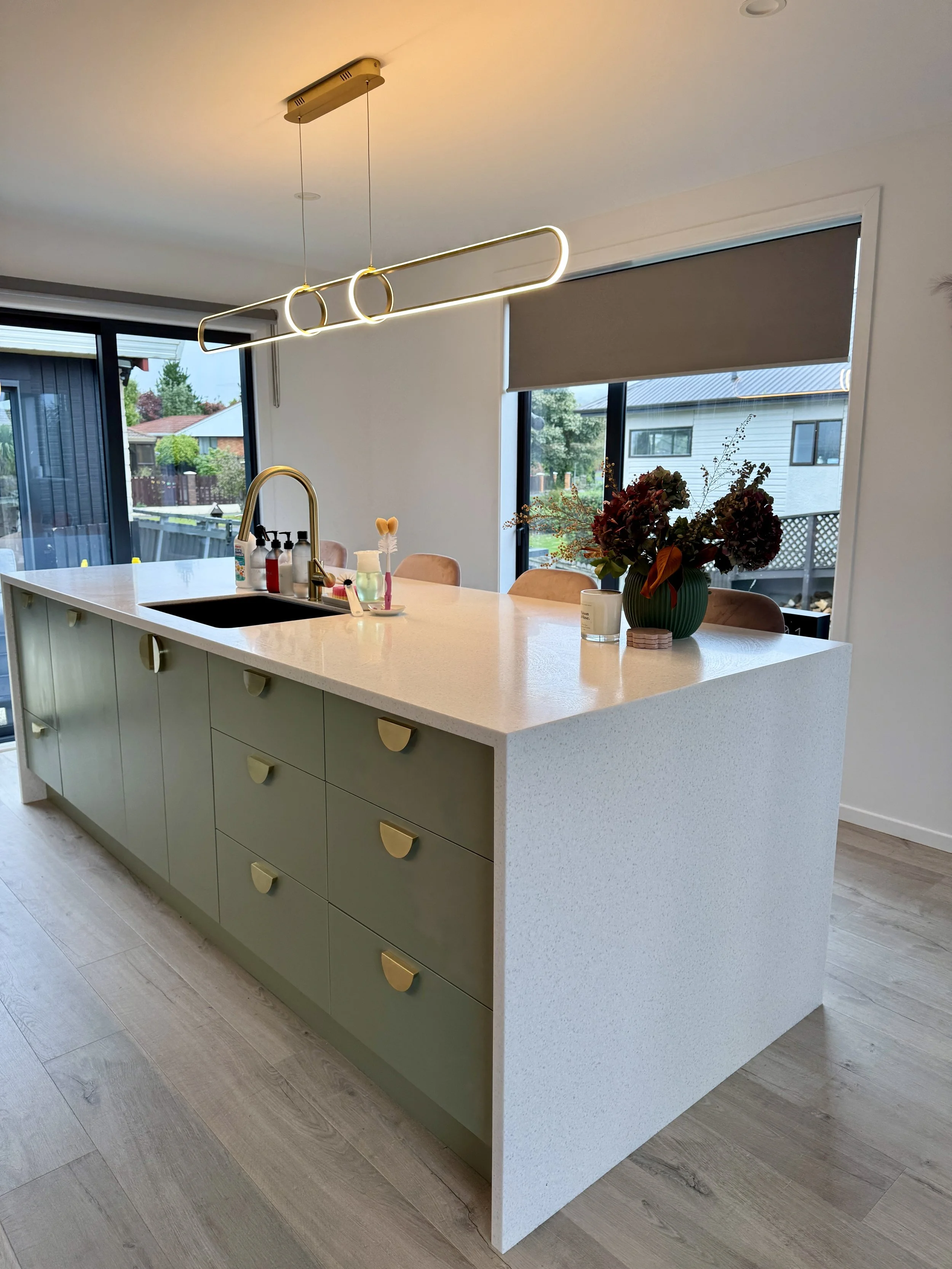 Modern kitchen island with beige drawers and a white countertop, with a dark sink, a gold faucet, and decorative items including a sunflower-shaped object, soap bottles, a green vase with flowers, and a candle. Large windows with blinds and a view of