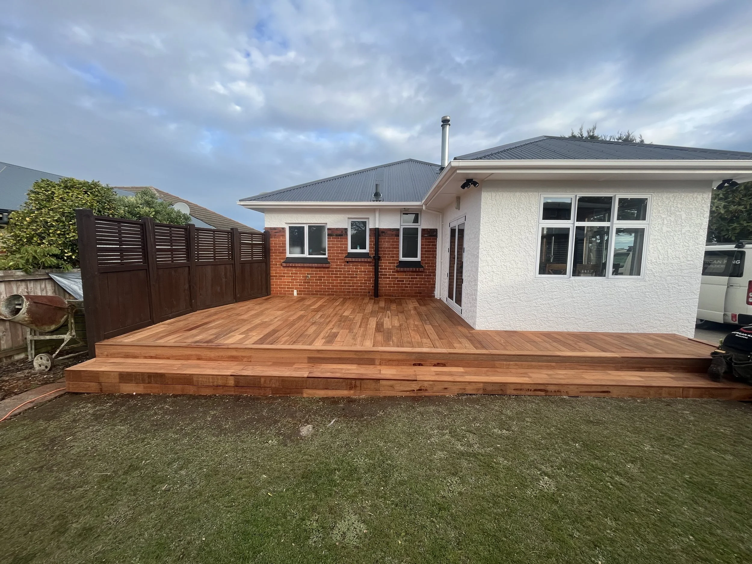A backyard with a newly installed wooden deck, a white house with brick accents, a high wooden fence, and an overcast sky.