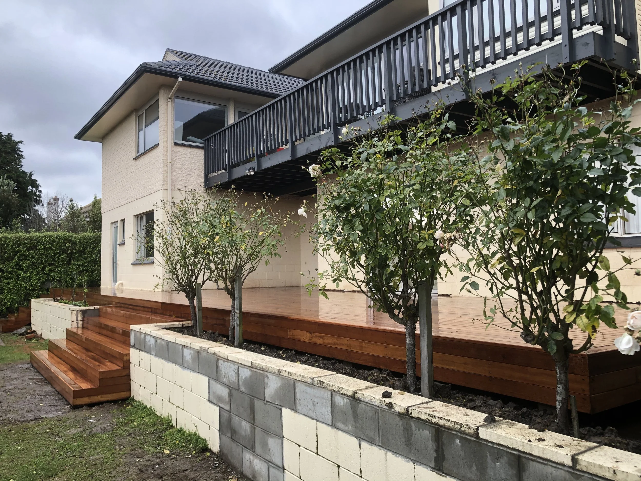 A residential house with a beige brick exterior and a balcony with black railing. There are steps leading up to a wooden deck outside, and there are five small trees or bushes in front of the house. The sky is overcast.