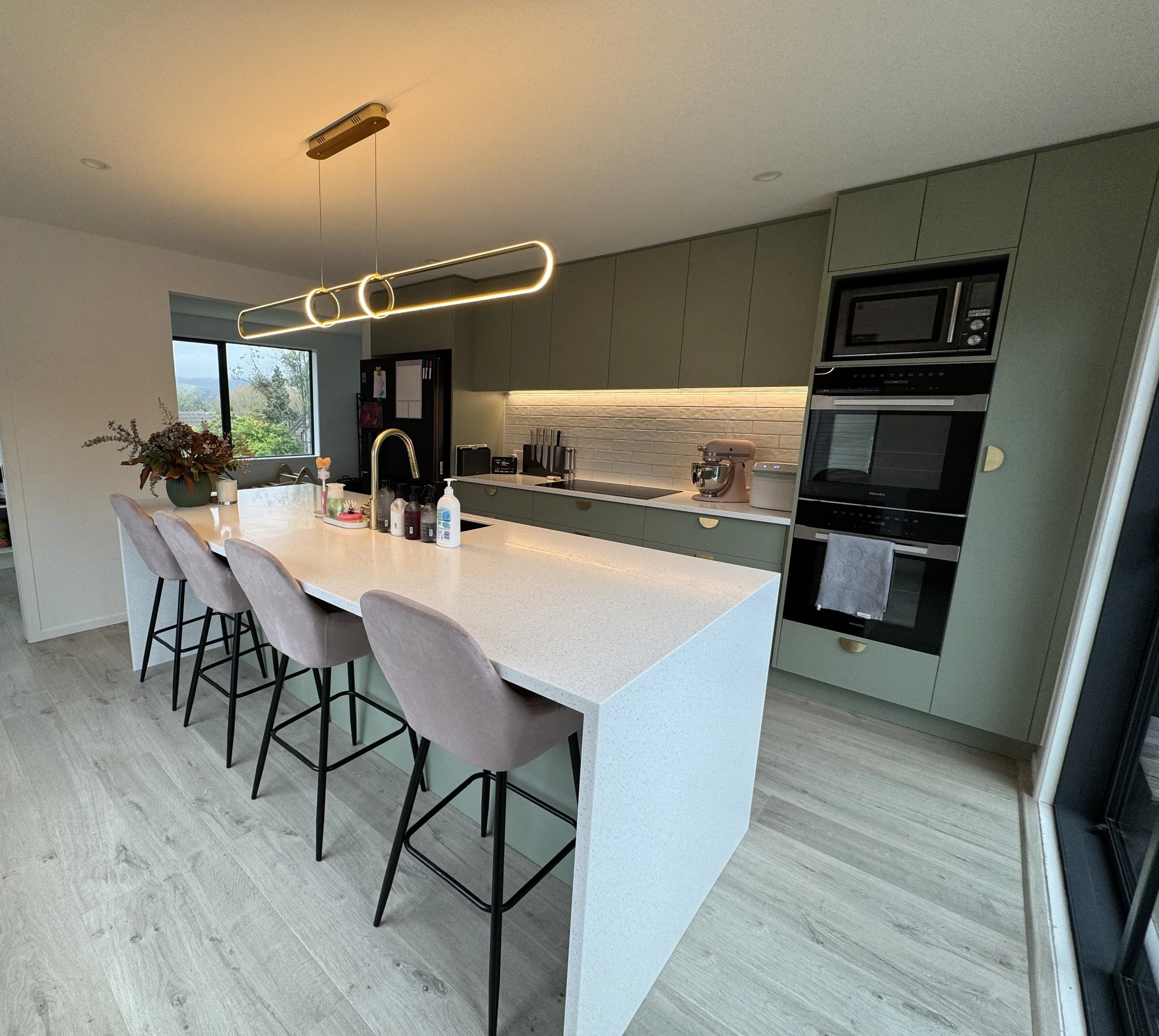 Contemporary kitchen with a white island countertop, pink upholstered bar stools, green cabinetry, and modern lighting fixtures.