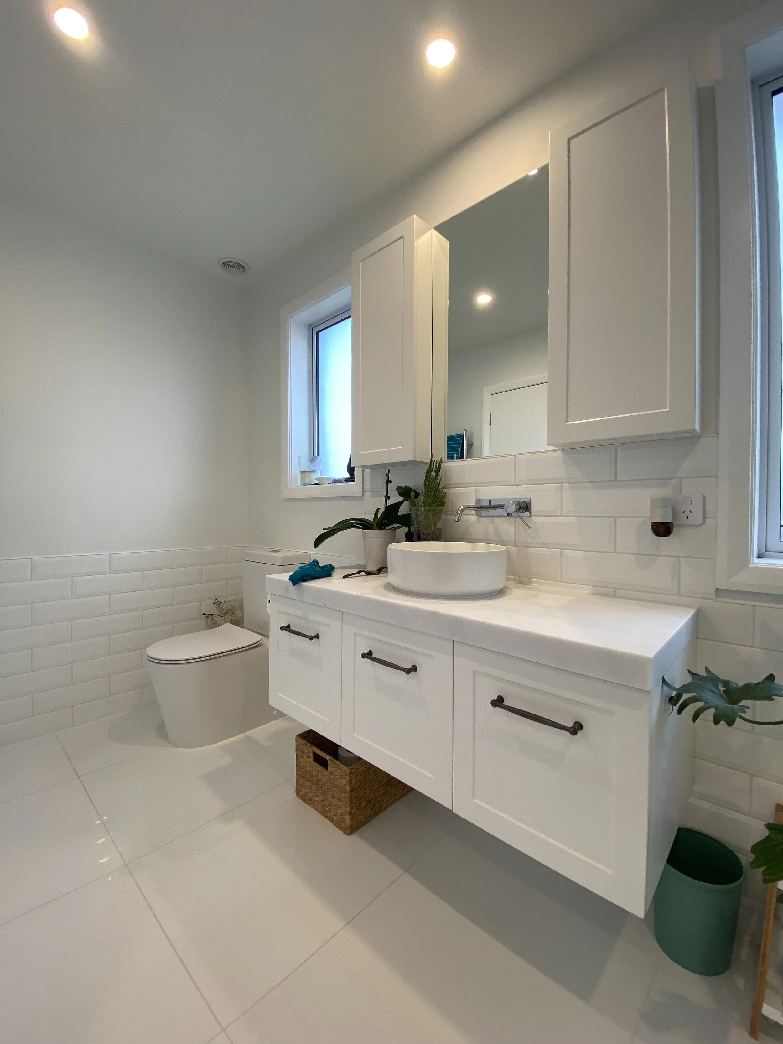 Modern bathroom with white cabinets, a white vessel sink, and a mirror. There are two small windows with frosted glass, a potted plant on the counter, and a woven basket below. The floor and walls are tiled in white with some decorative tile accents.