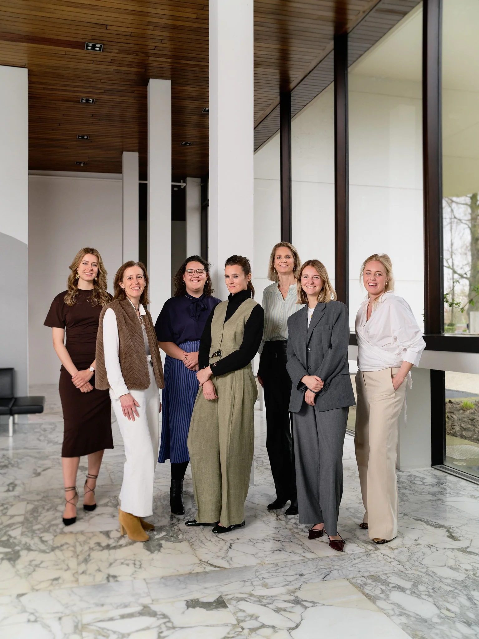 A group of seven women standing in a modern, bright room with large windows, wearing professional attire, smiling, and posing for the photo.
