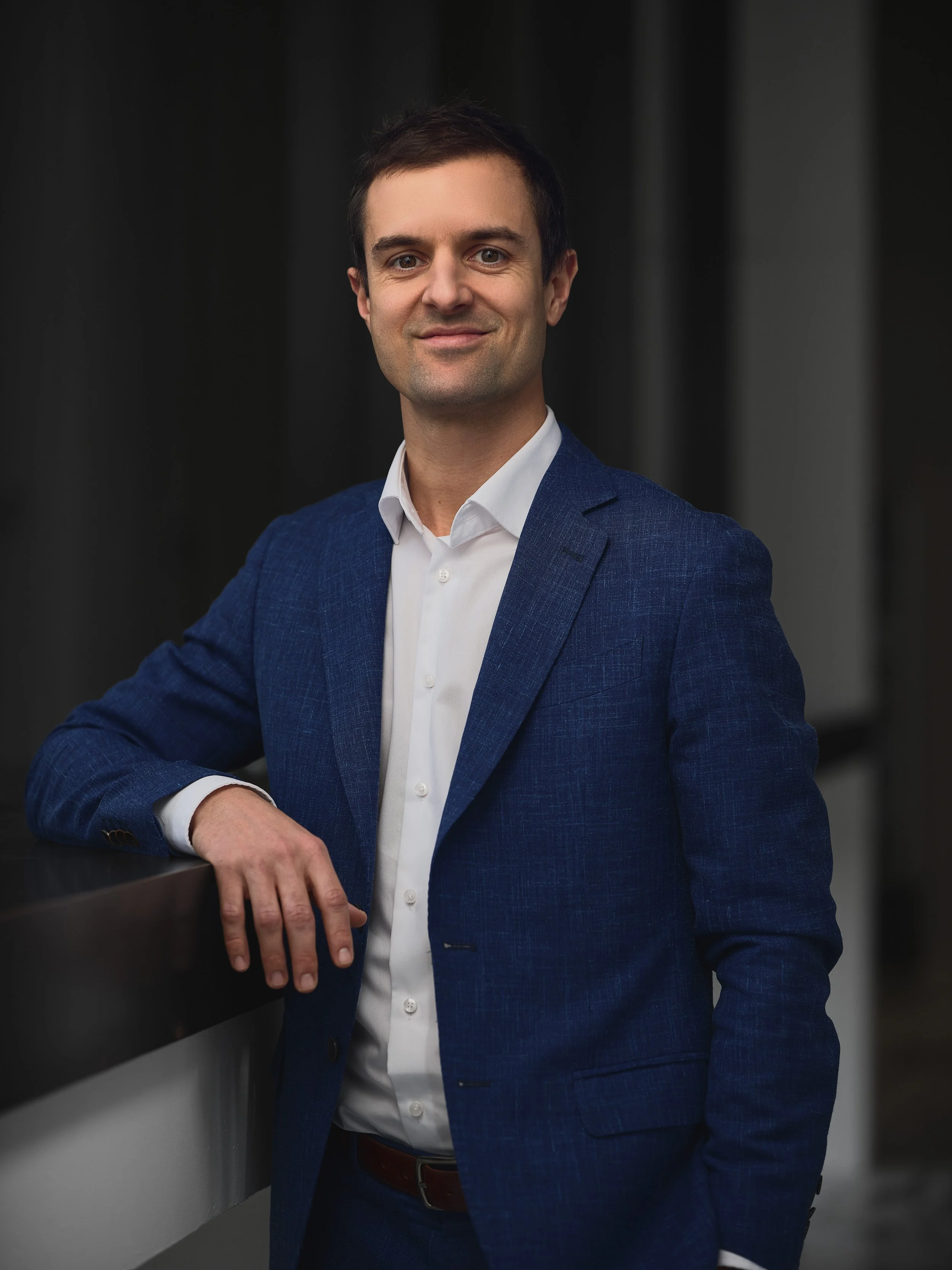 A man in a blue blazer and white shirt stands with his arm resting on a ledge, smiling at the camera against a dark, blurred background.