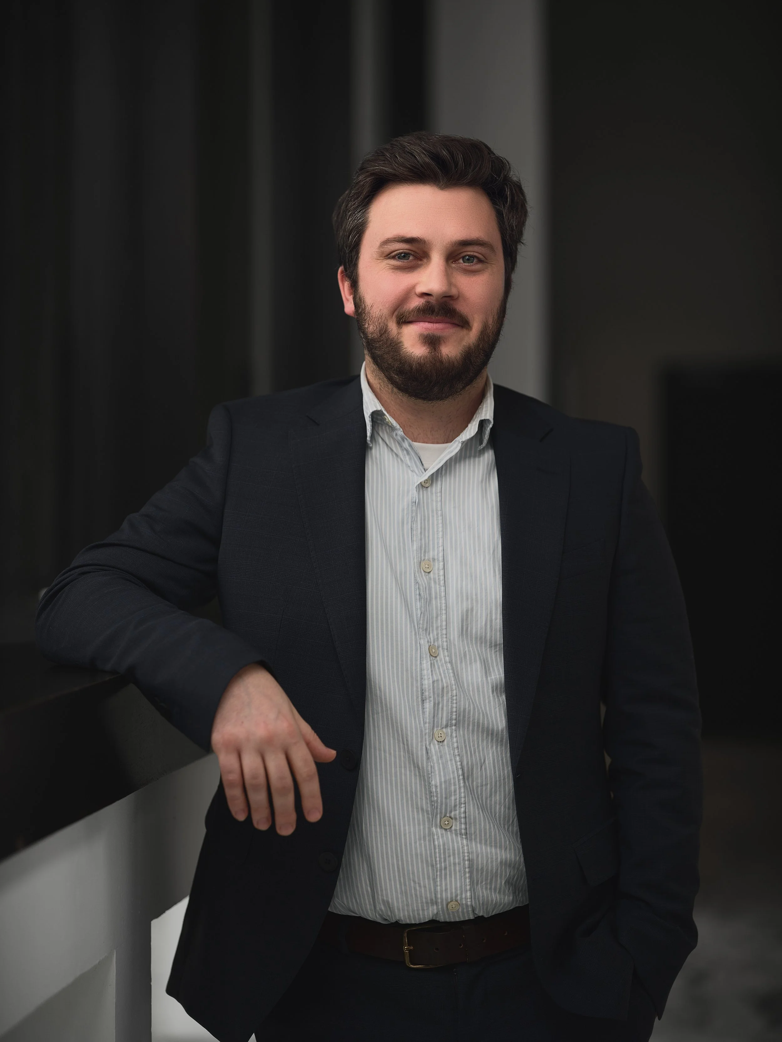 A man with dark hair and a beard, wearing a dark suit and a light striped shirt, leaning against a black railing in an indoor setting.