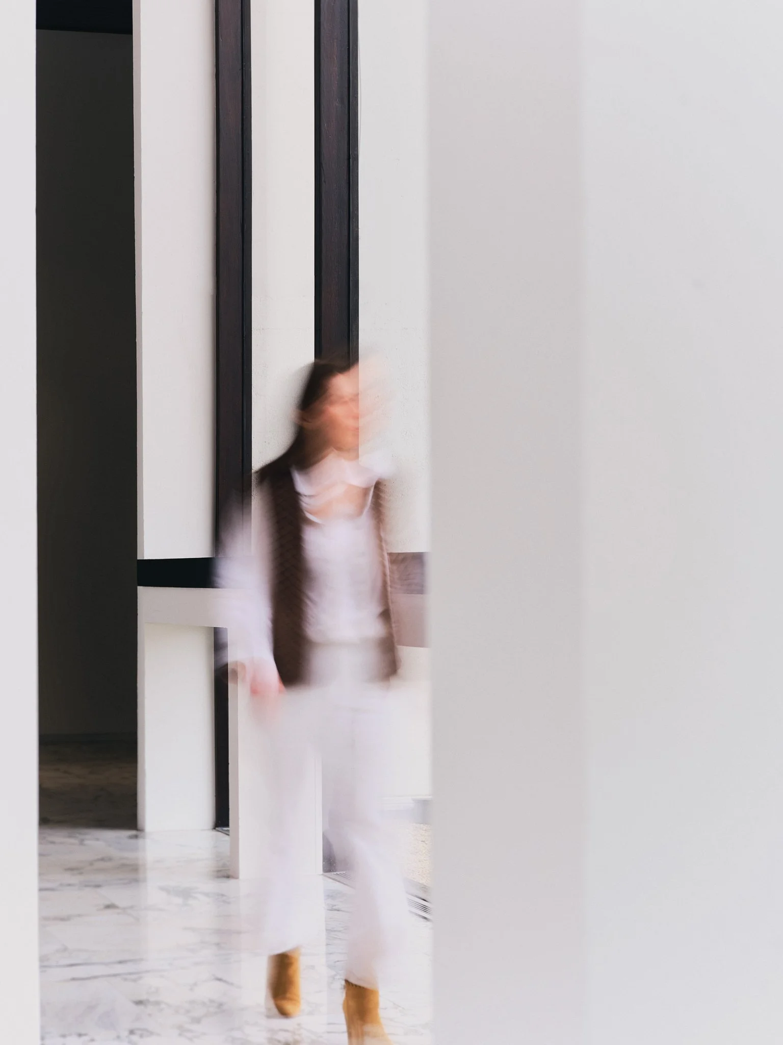 A woman wearing a white dress, brown cardigan, and tan boots walking past a mirror with a reflection showing her face and upper body, in a modern, minimalistic interior space with white walls and marble flooring.