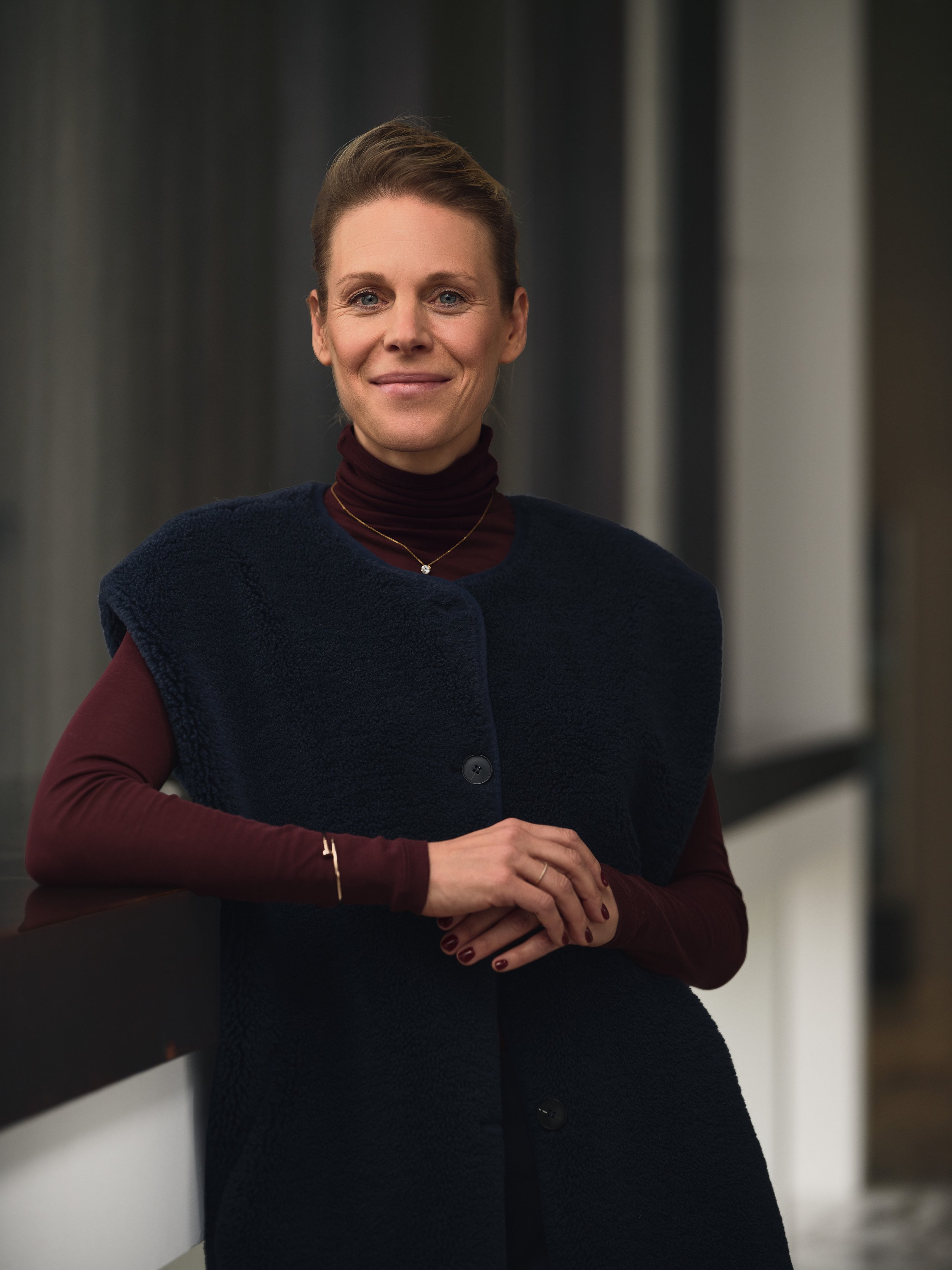 A woman with short hair smiling, standing indoors in front of a dark curtain.