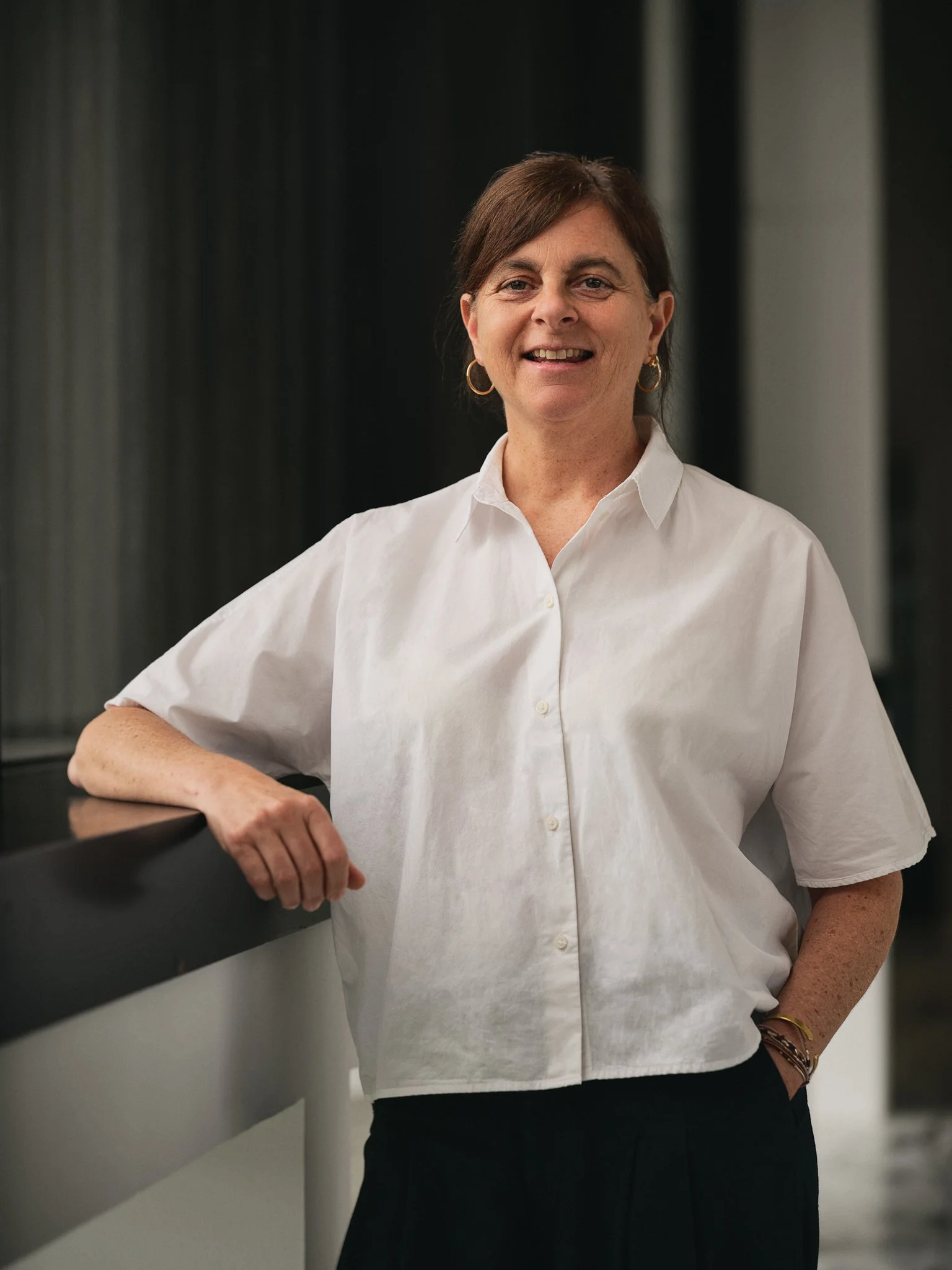 A woman standing indoors, wearing a white short-sleeved button-up shirt, gold hoop earrings, and a bracelet, smiling with one arm resting on a ledge.