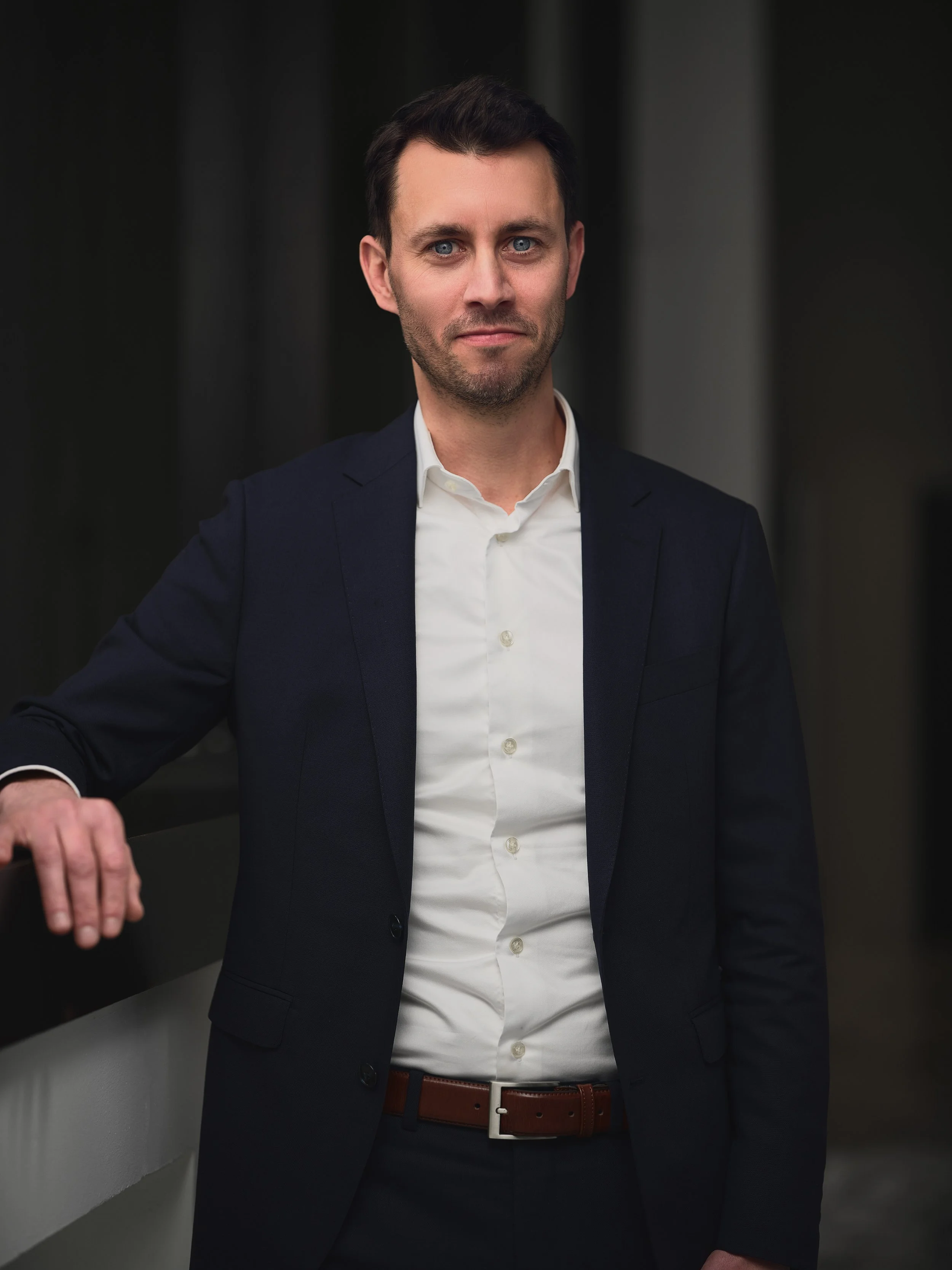 A man in a dark suit and white shirt standing indoors, with his left arm resting on a surface.