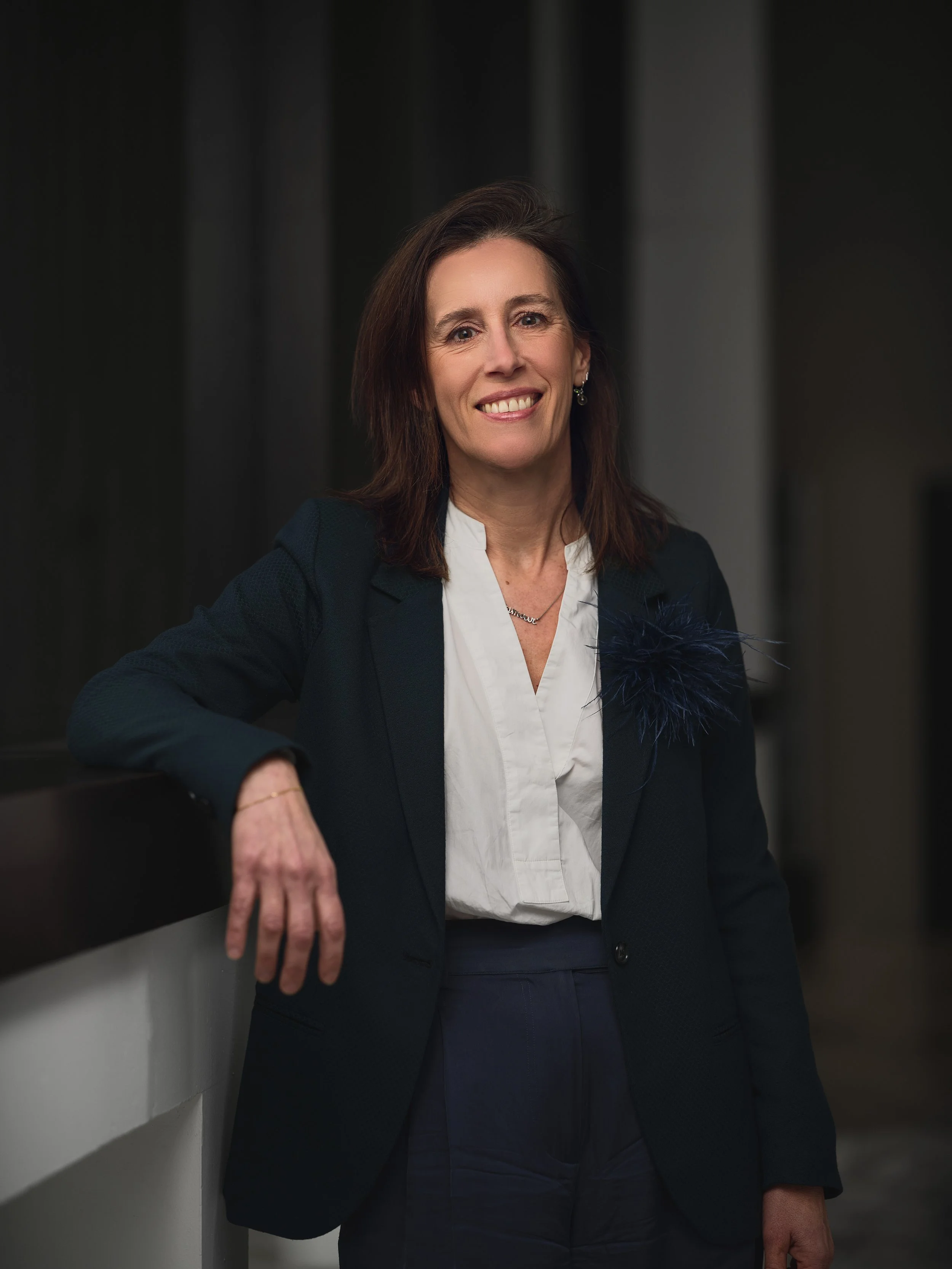 A woman with brown hair wearing a black blazer, white blouse, and navy pants, smiling and leaning against a gray wall.