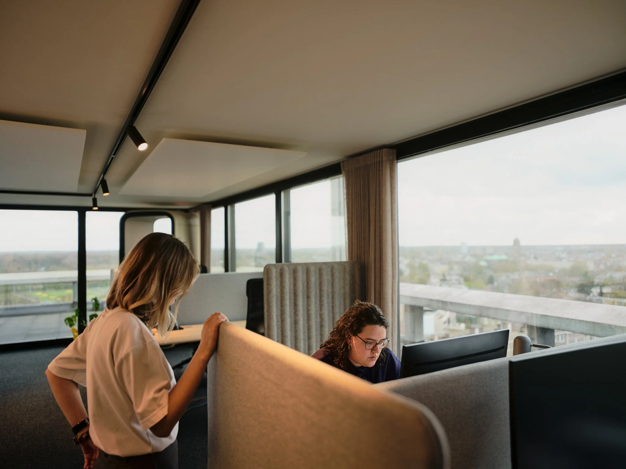 Two women working on computers in a modern office with large windows and a city view.