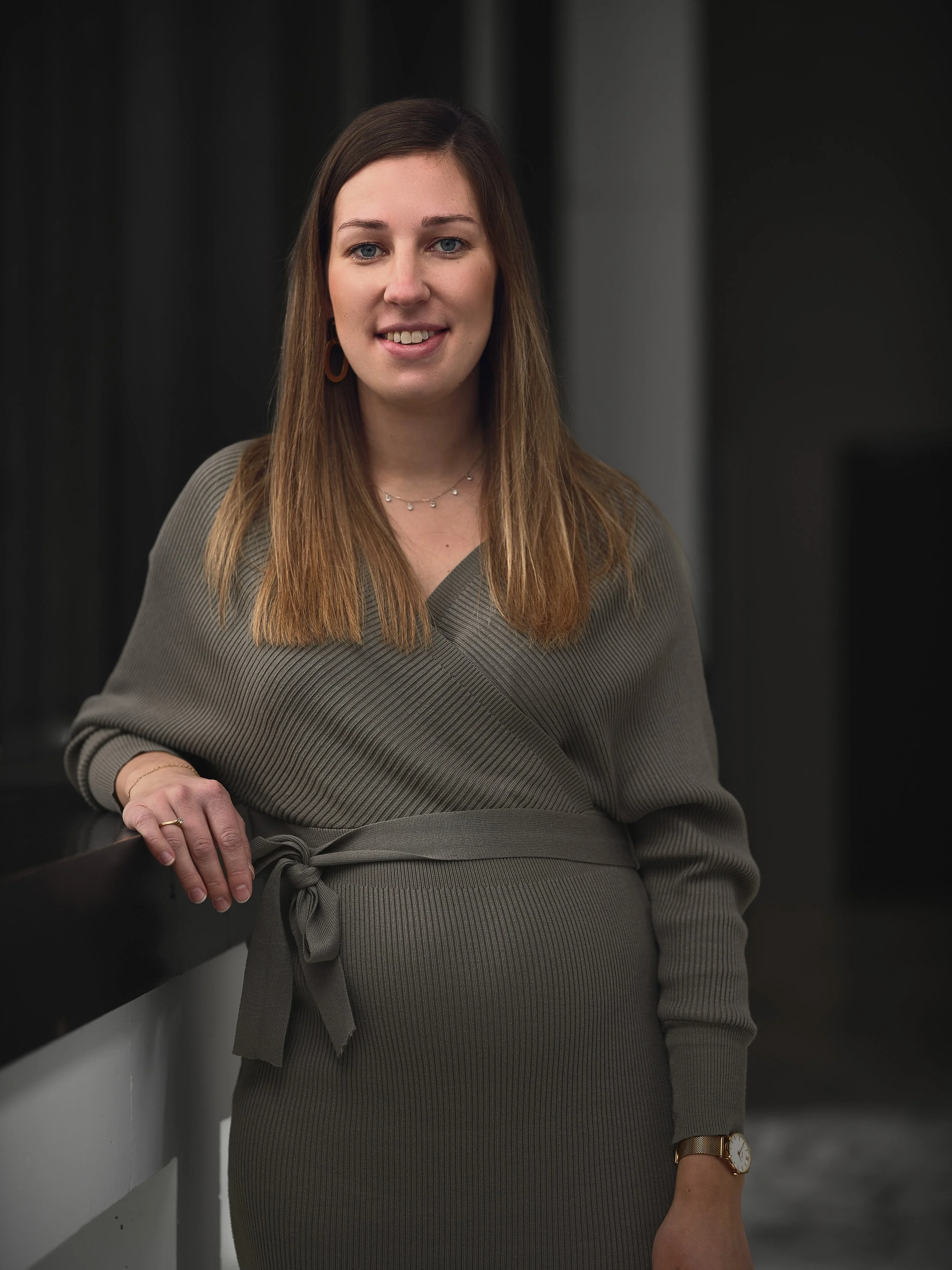 A woman with long brown hair wearing a taupe wrap dress, gold watch, and jewelry standing in an indoor space near a black railing.