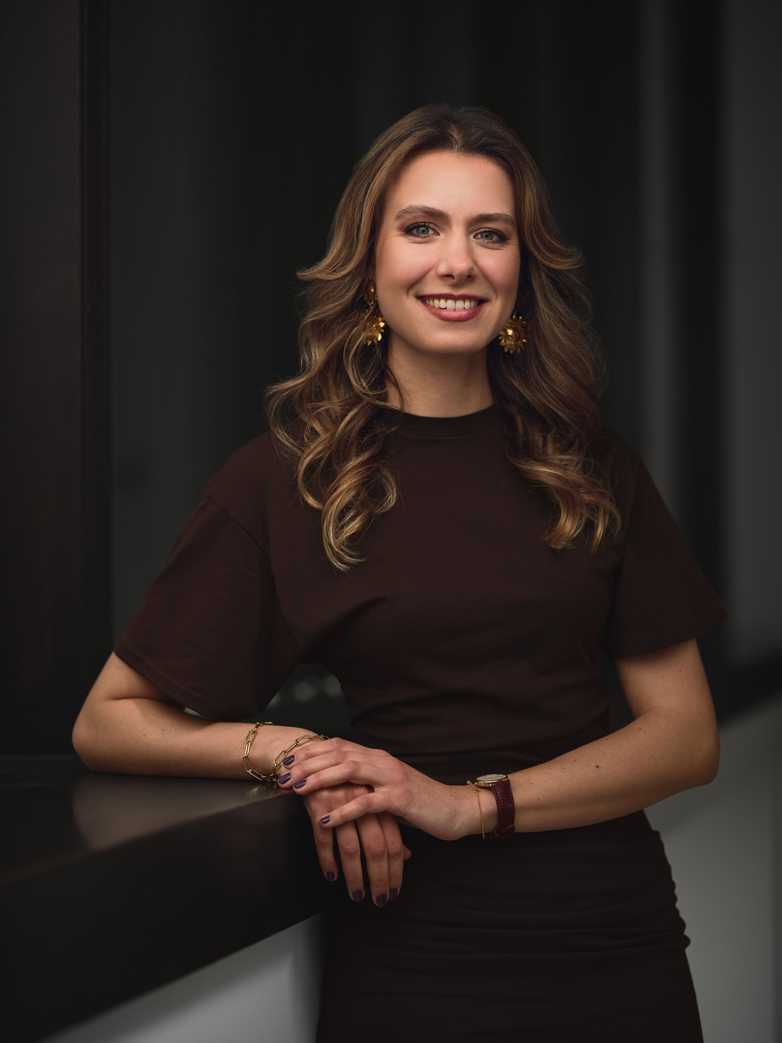 A woman with wavy brown hair wearing a dark brown dress, gold earrings, and bracelets, standing against a black background with her arms resting on a dark surface.
