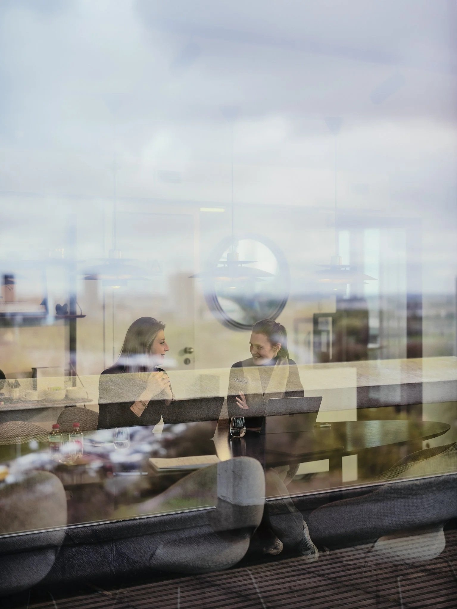 Two women are sitting at a table inside a cafe or restaurant, engaged in conversation. The scene is viewed through a glass window, which reflects the outside landscape.