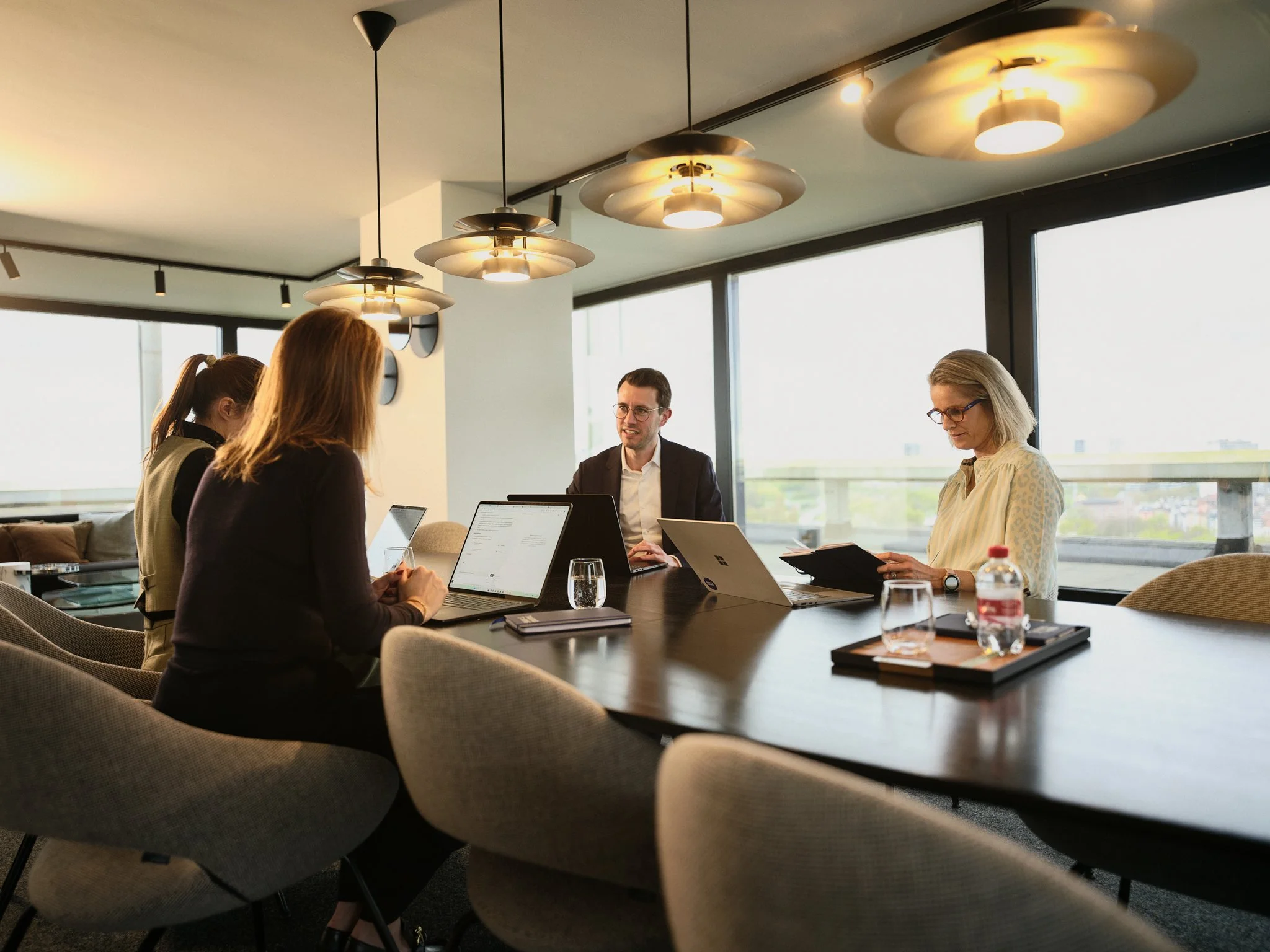 Four people sitting around a conference table in a modern office, working on laptops and tablets, with large windows and contemporary lighting fixtures overhead.
