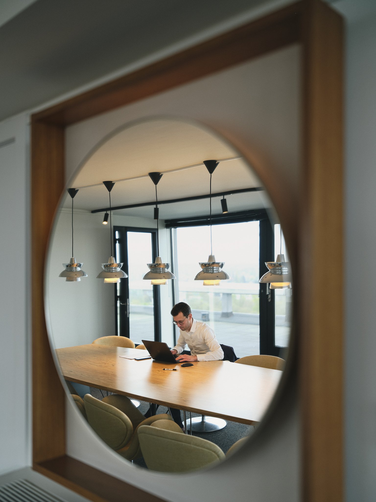 A man working on a laptop at a conference room table, seen through a decorative oval mirror.