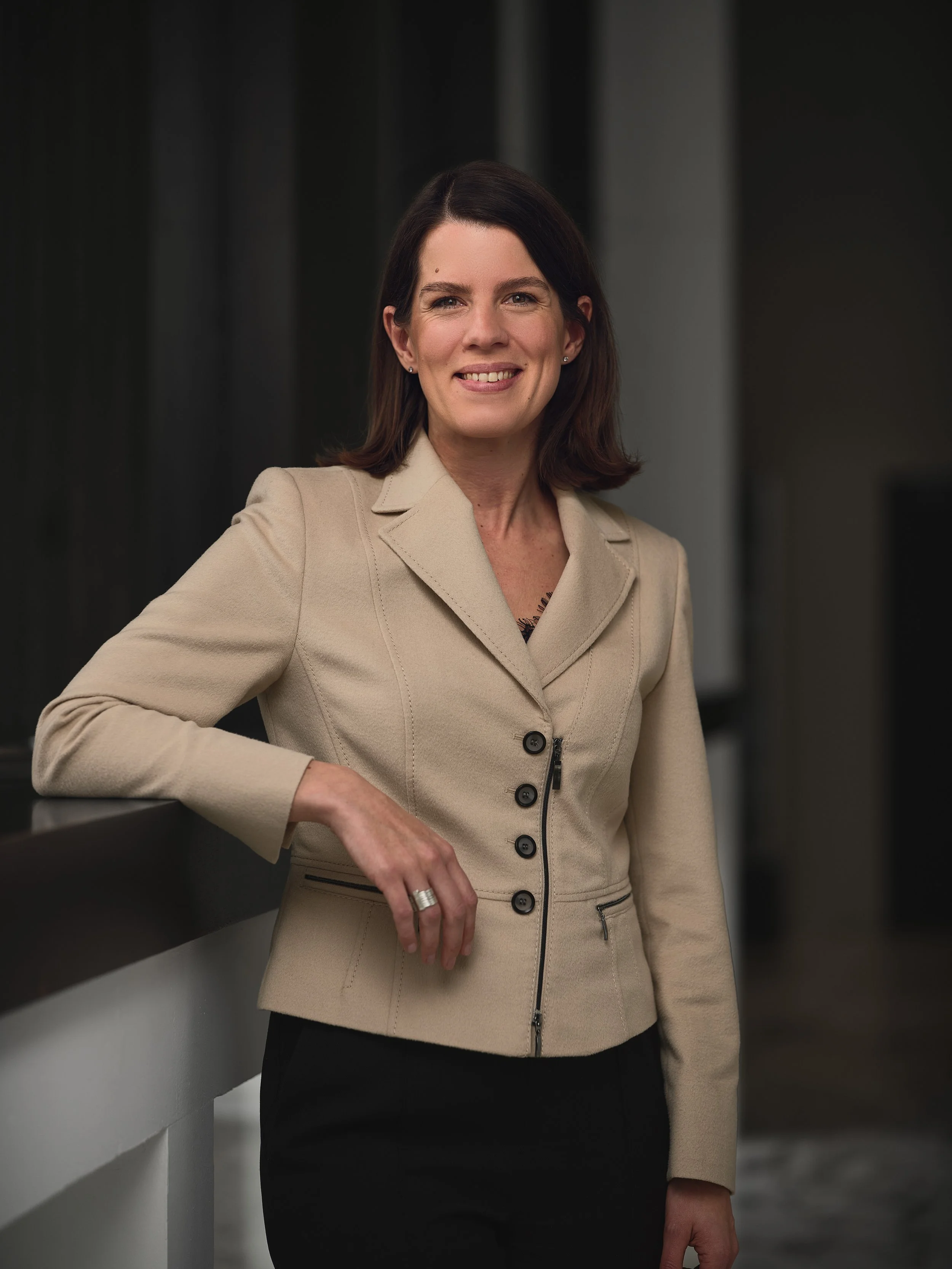 A woman with dark brown hair and a beige blazer smiling while leaning against a dark-colored railing in an indoor setting.