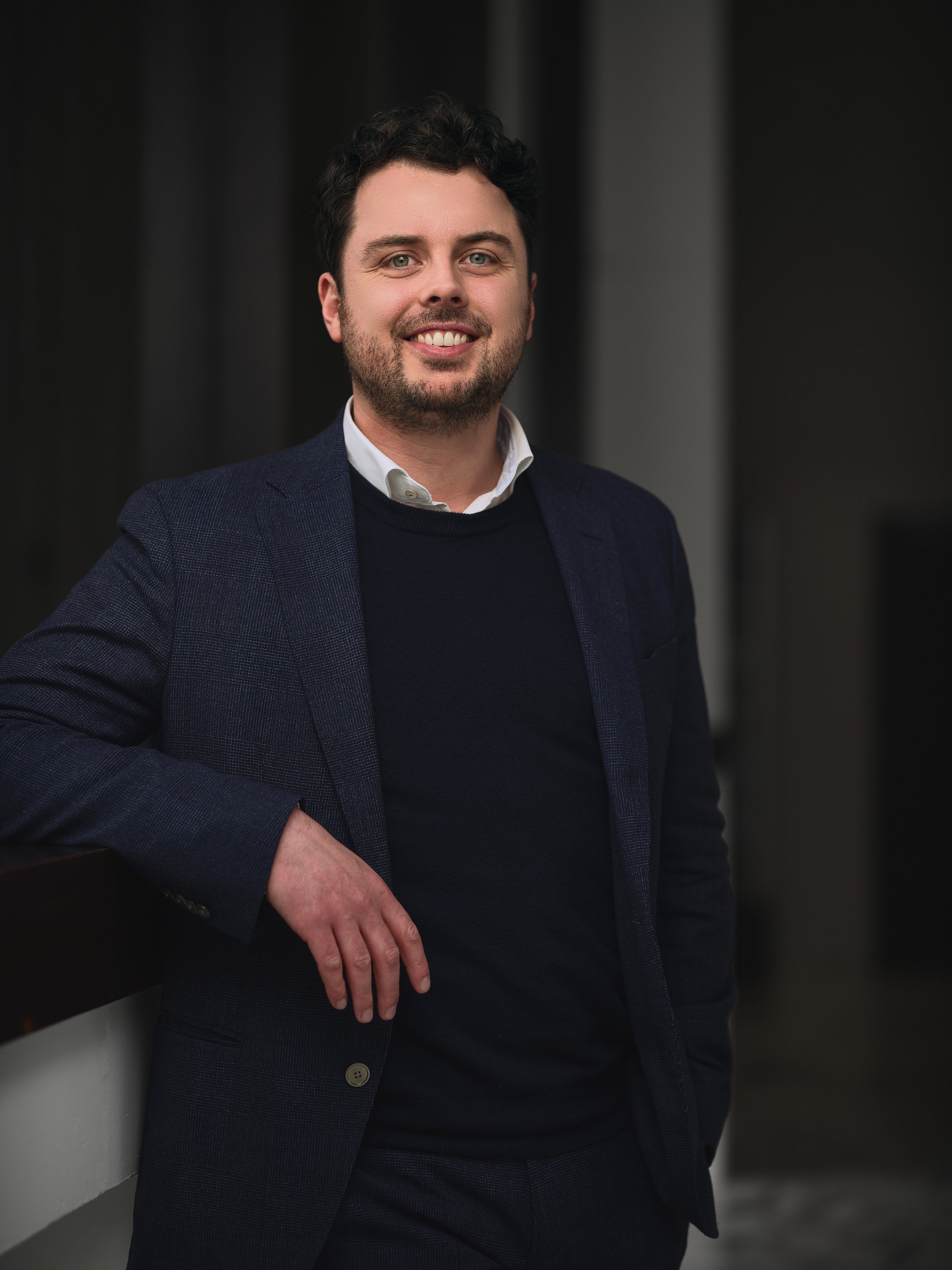 A man with dark, curly hair and light skin tone wearing a dark blue blazer, white shirt, and dark sweater, standing in a professional setting with a dark background.