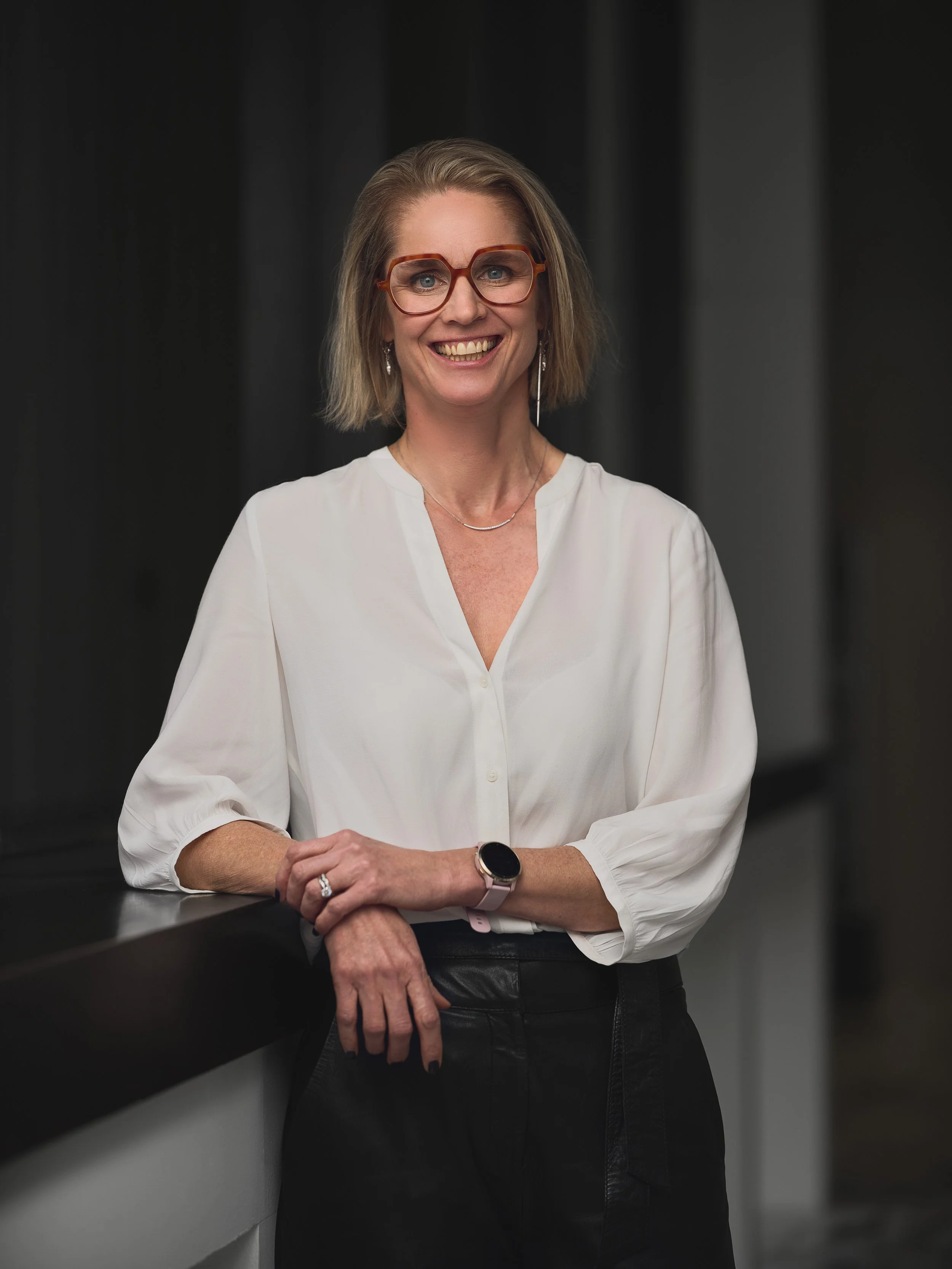 A woman with short blonde hair wearing glasses and a white blouse, smiling and leaning on a dark surface with her left hand resting on her right arm, in an indoor setting with dark curtains in the background.