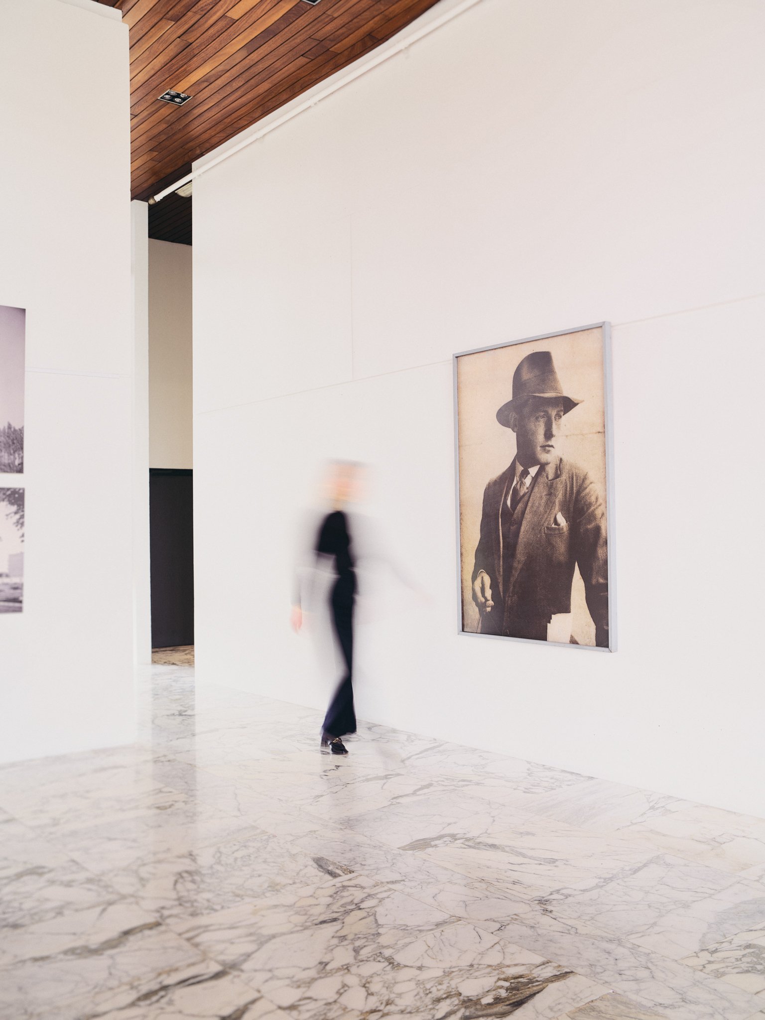A person walking past a black and white portrait of a man in a hat at an art gallery with white walls and marble floor.