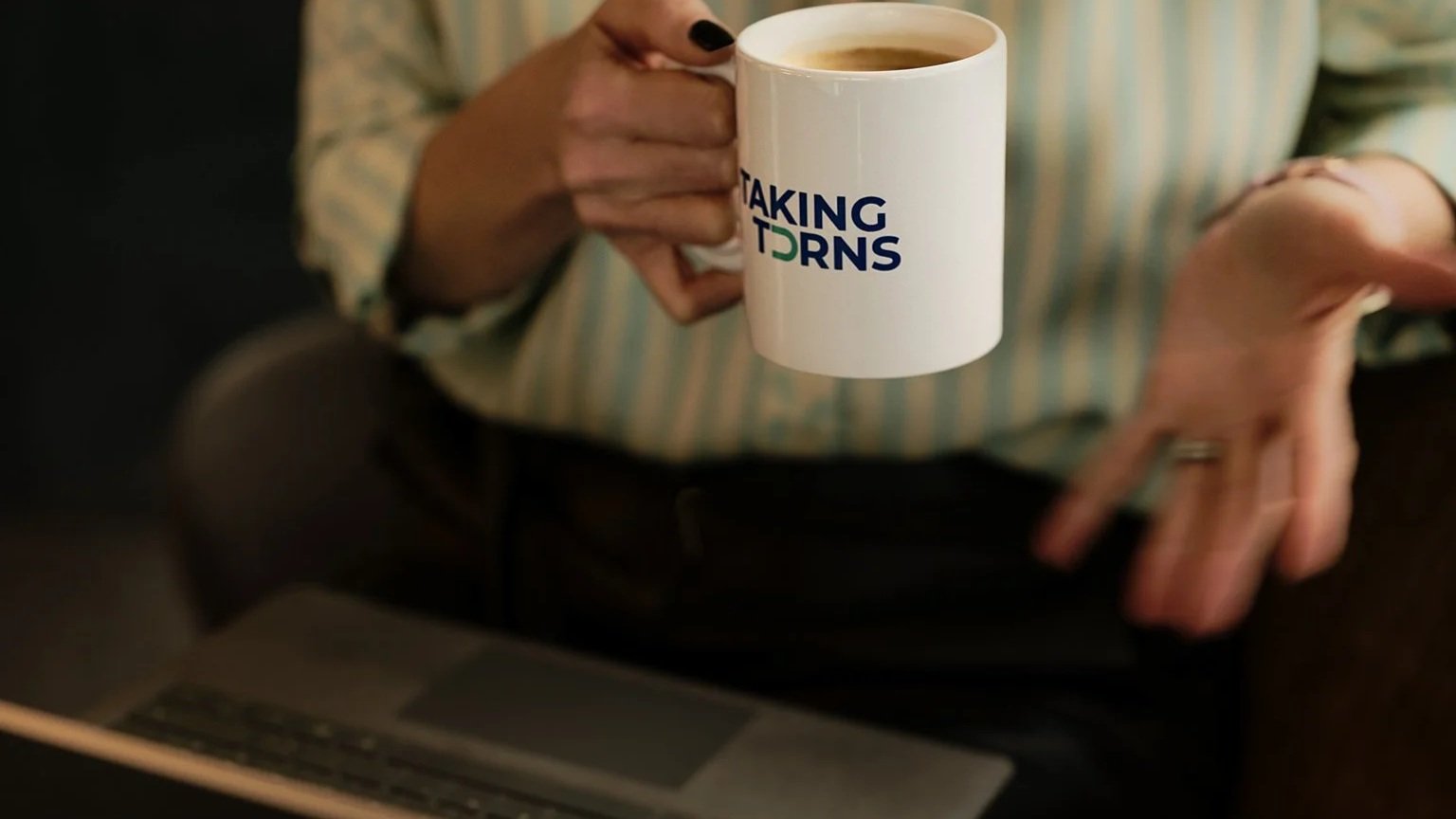 Person holding a white coffee mug with the text "TAKING TURNS" printed on it, while sitting at a table with a laptop.