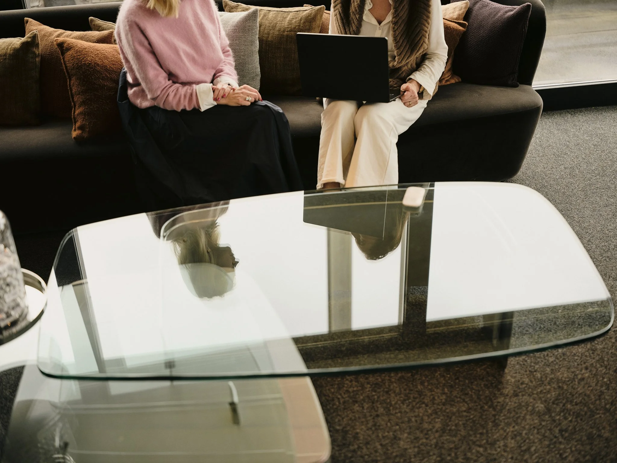 A coffee table in front of a glass window, with a reflection showing the top of a person's head and shoulder. The background shows two women sitting on a dark couch, one wearing a pink sweater and the other a white shirt with a beige scarf, with colorful pillows behind them.