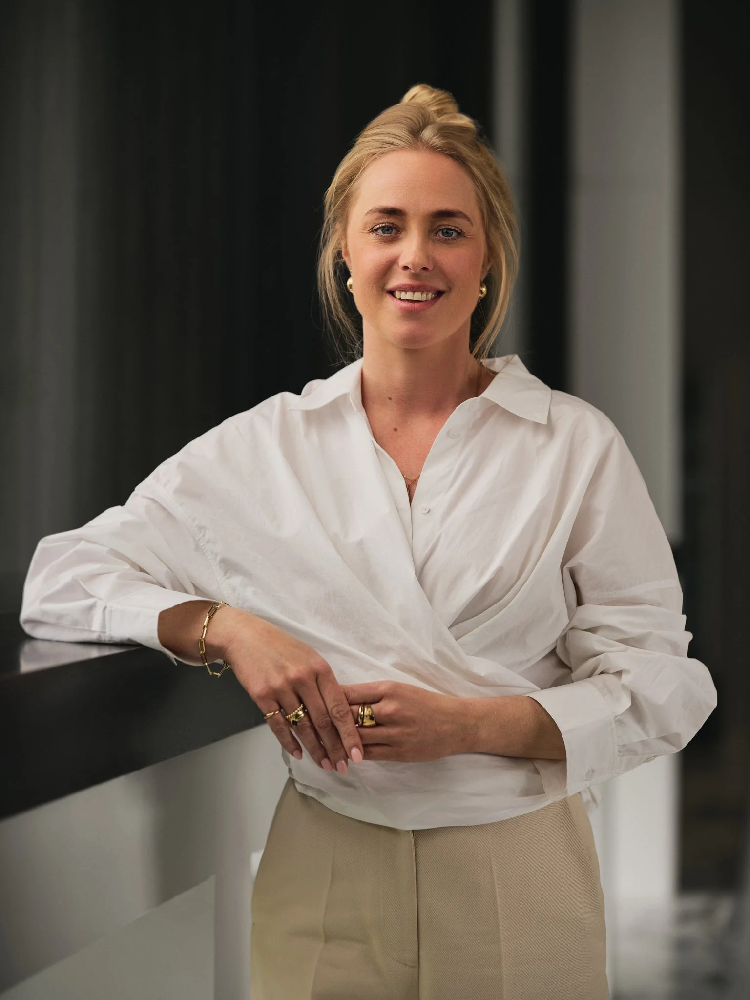 A woman dressed in a white blouse and beige pants, leaning on a black ledge with a dark background.