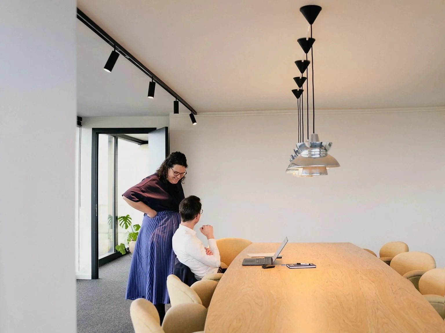 Two women in a conference room, one seated with a laptop and the other standing, engaging in conversation.