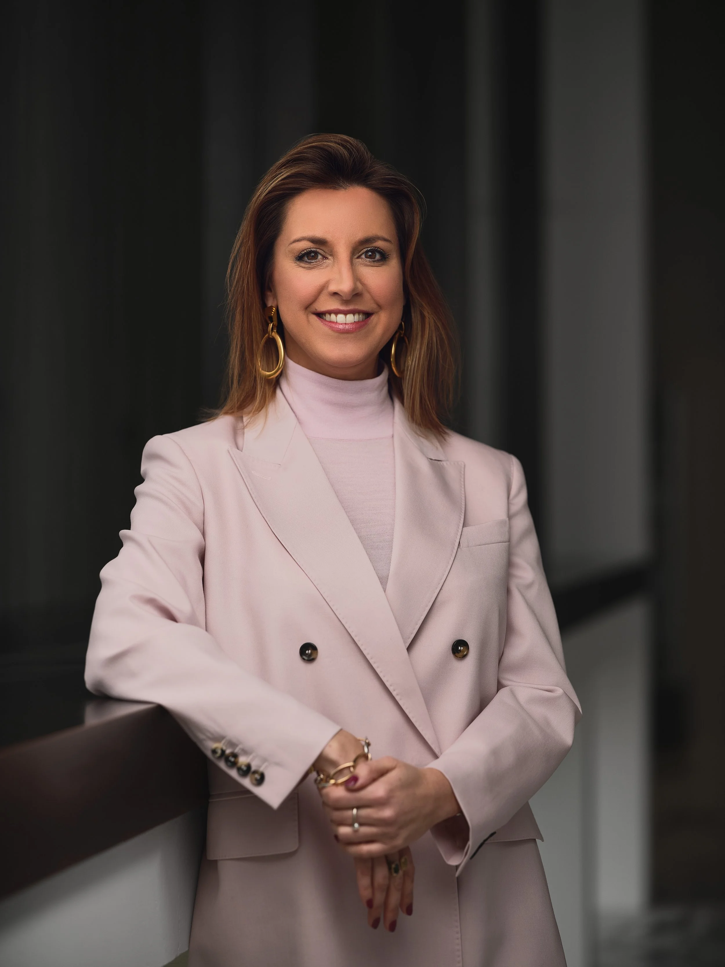 A woman with shoulder-length brown hair smiling, wearing a light pink blazer with gold buttons, gold earrings, and multiple rings, standing against a black background.