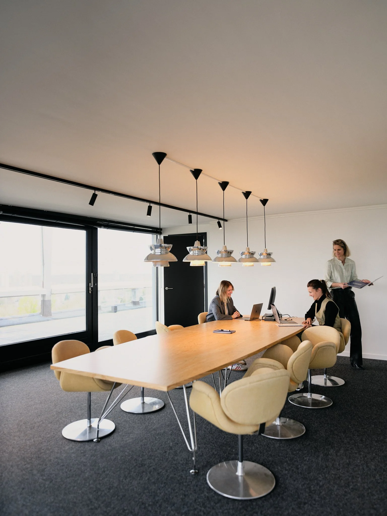 A modern conference room with a long wooden table, beige chairs, hanging light fixtures, large windows, and three women working at laptops.