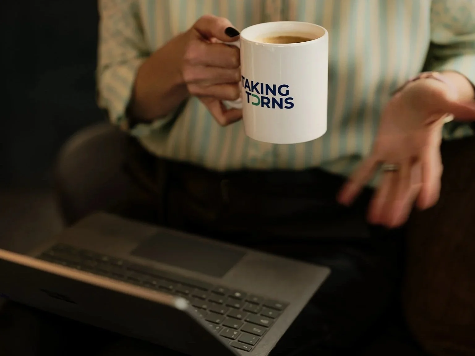 Person holding a white coffee mug with blue and green text that says 'Taking Turns' in front of a laptop keyboard, wearing a striped shirt.