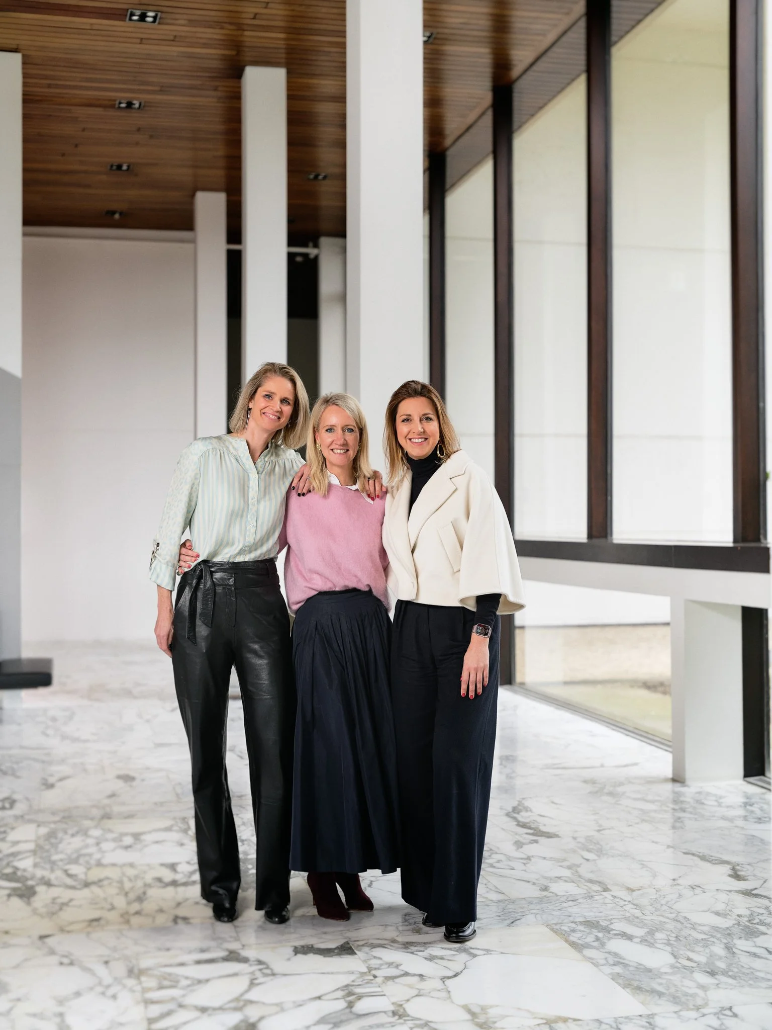 Three women standing together in a modern building with marble flooring and large windows, smiling for a photo.