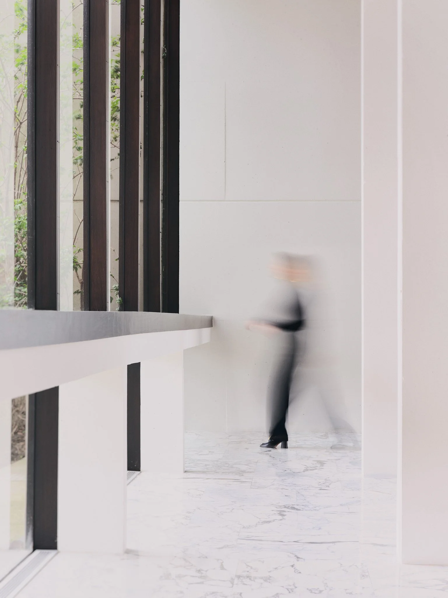 A blurred woman in black heels and a black outfit walking through a modern interior space with white marble floor, white wall, and wooden beams beside a window showing green trees outside.