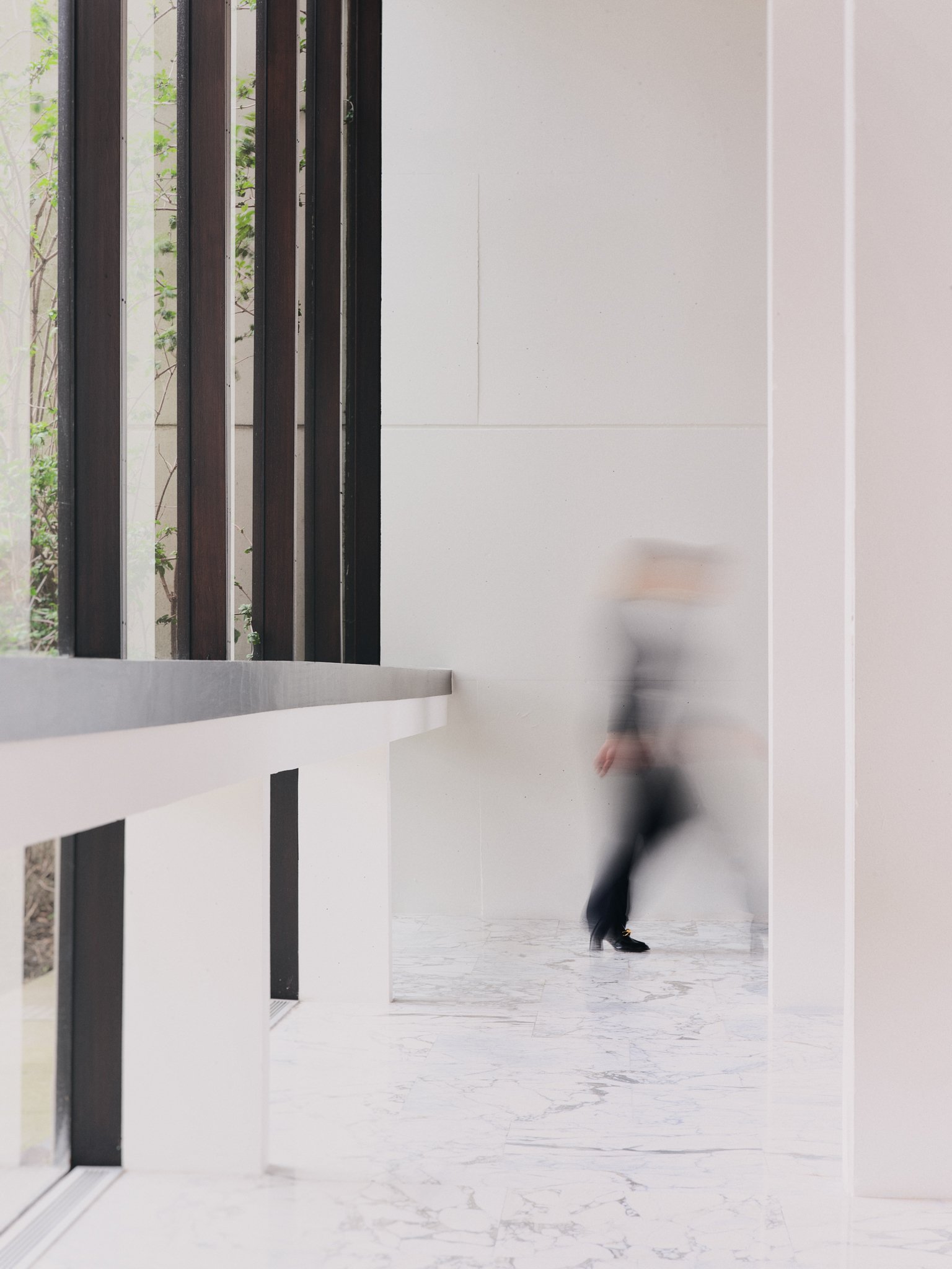 Blurred person walking in a room with white walls and marble flooring, seen through a window with black frames.