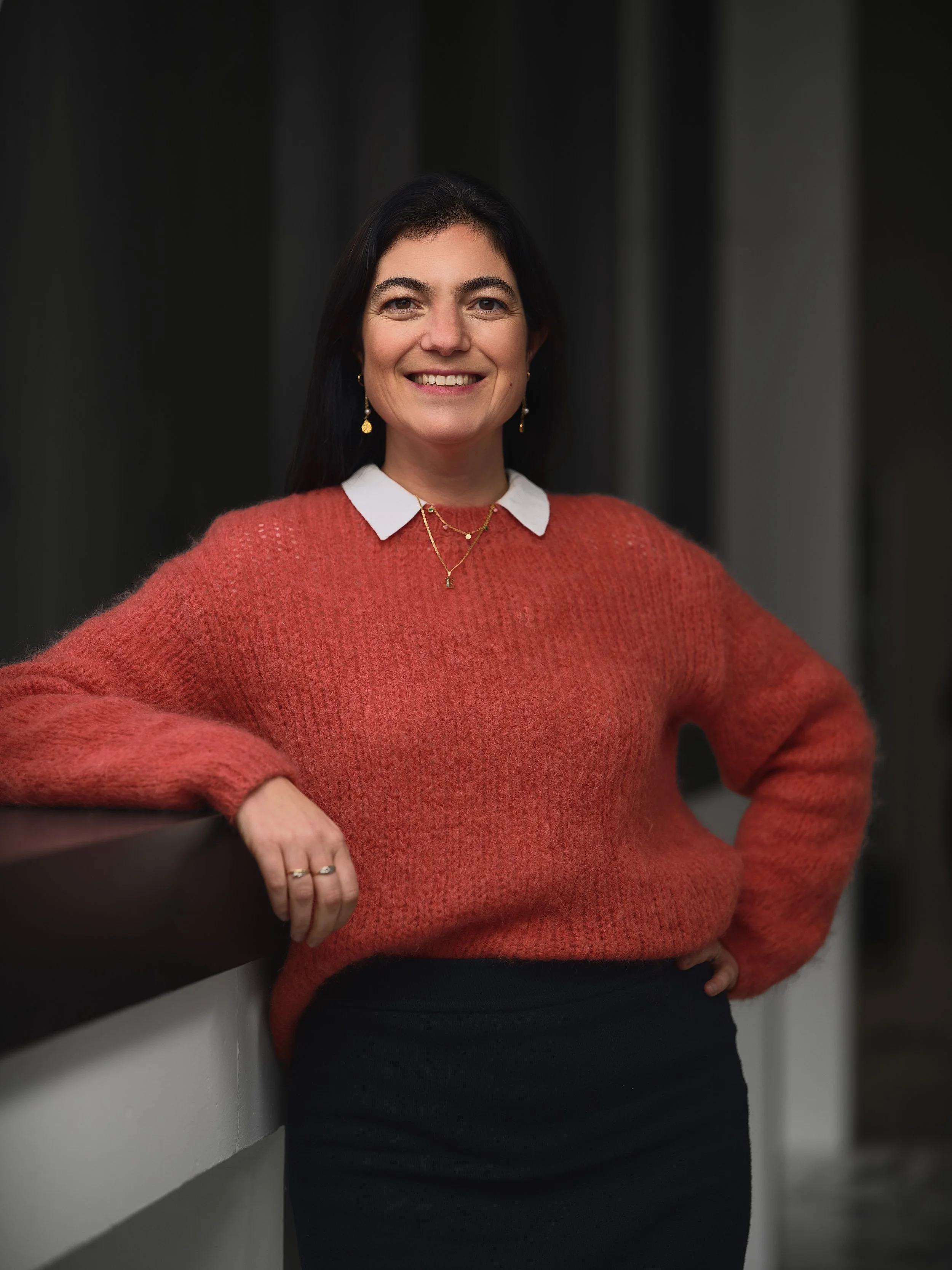Woman with dark hair smiling, wearing a red sweater over a white collared shirt, gold jewelry, standing against a dark background.