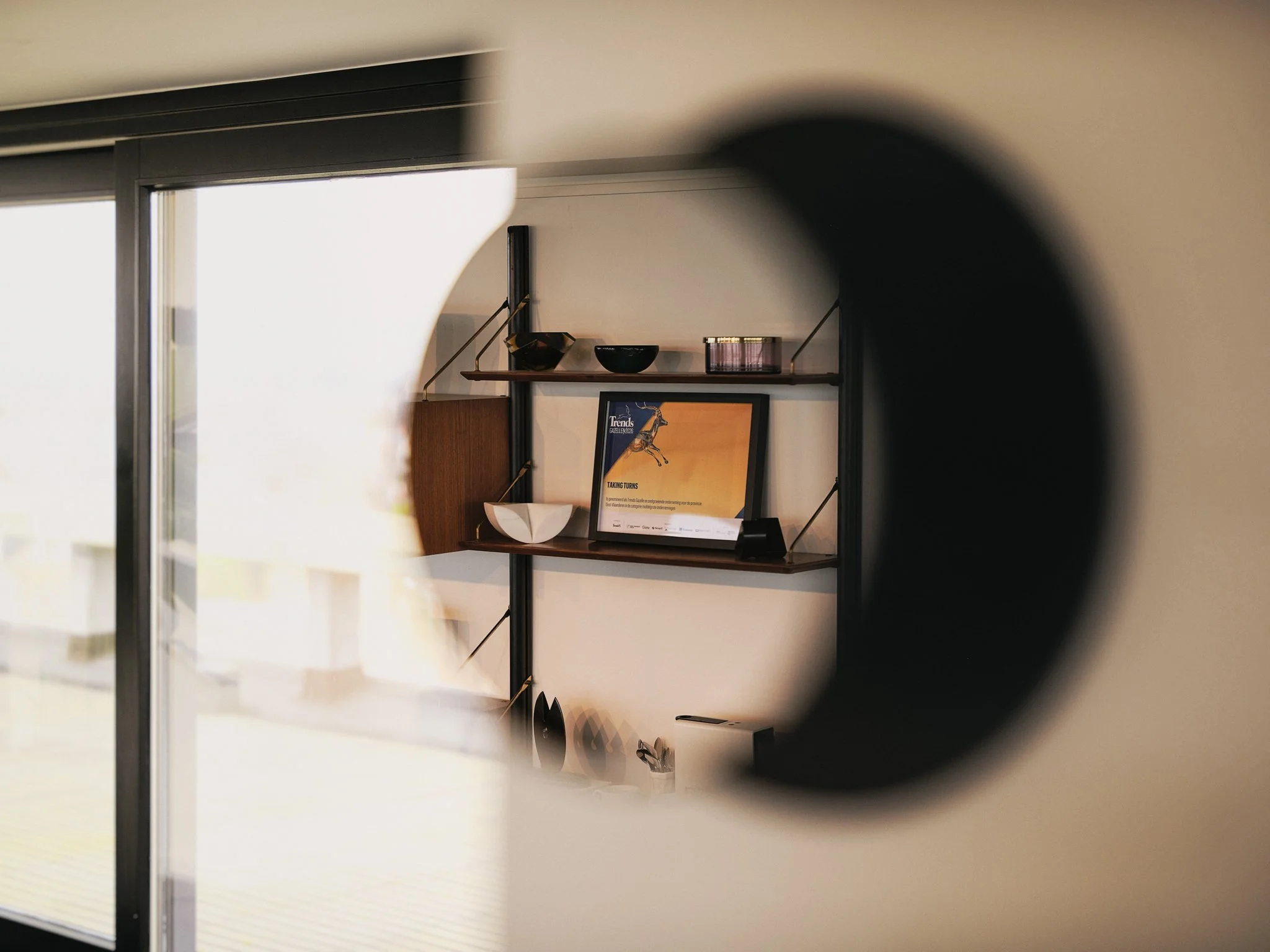 View of a shelf with decorative items including bowls, a framed picture, and a sculpture, seen through a circular opening in a wall or partition.