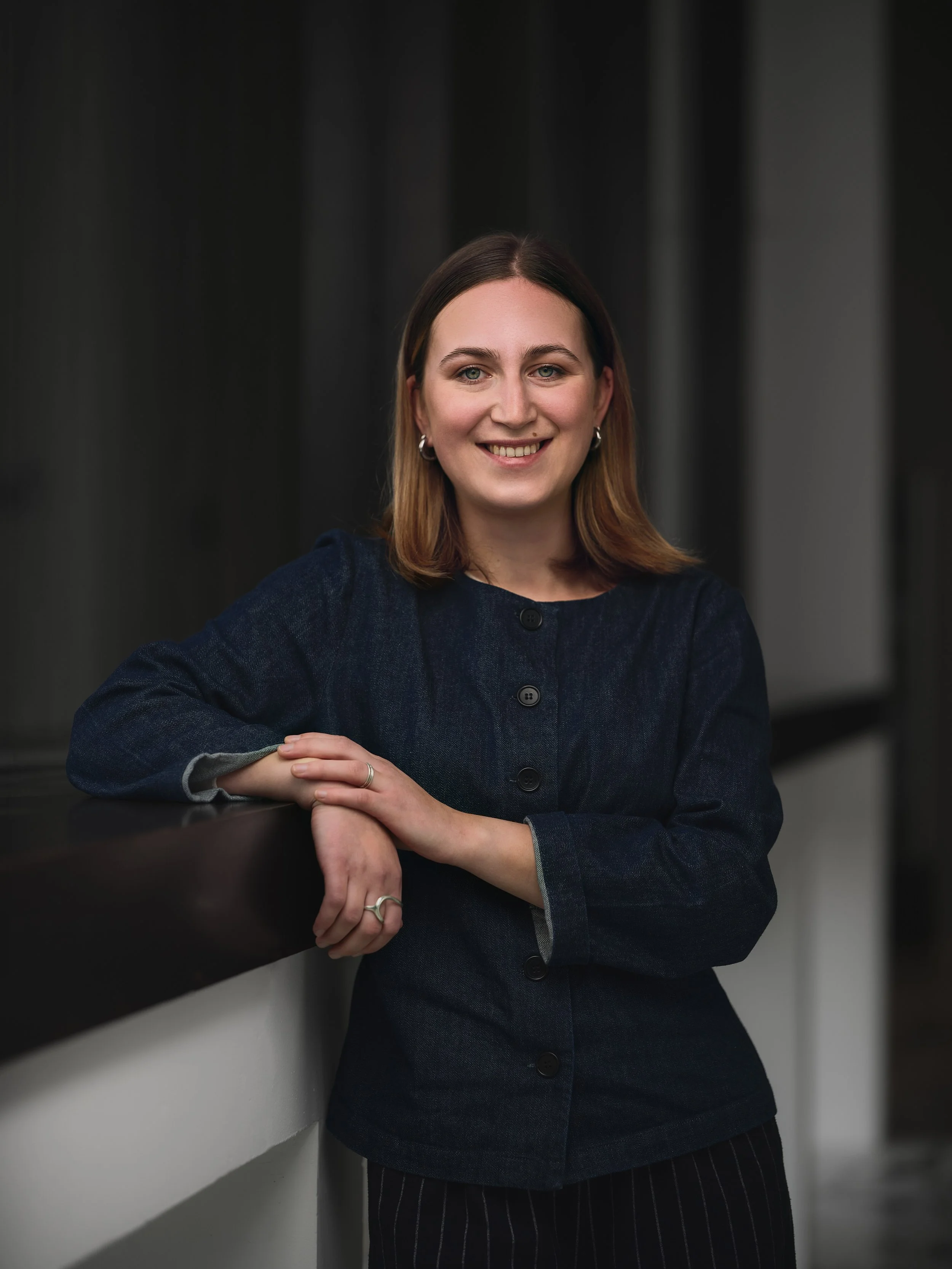 A smiling woman with shoulder-length brown hair, wearing a dark navy buttoned blazer and striped pants, leaning against a black surface in front of a dark curtain background.