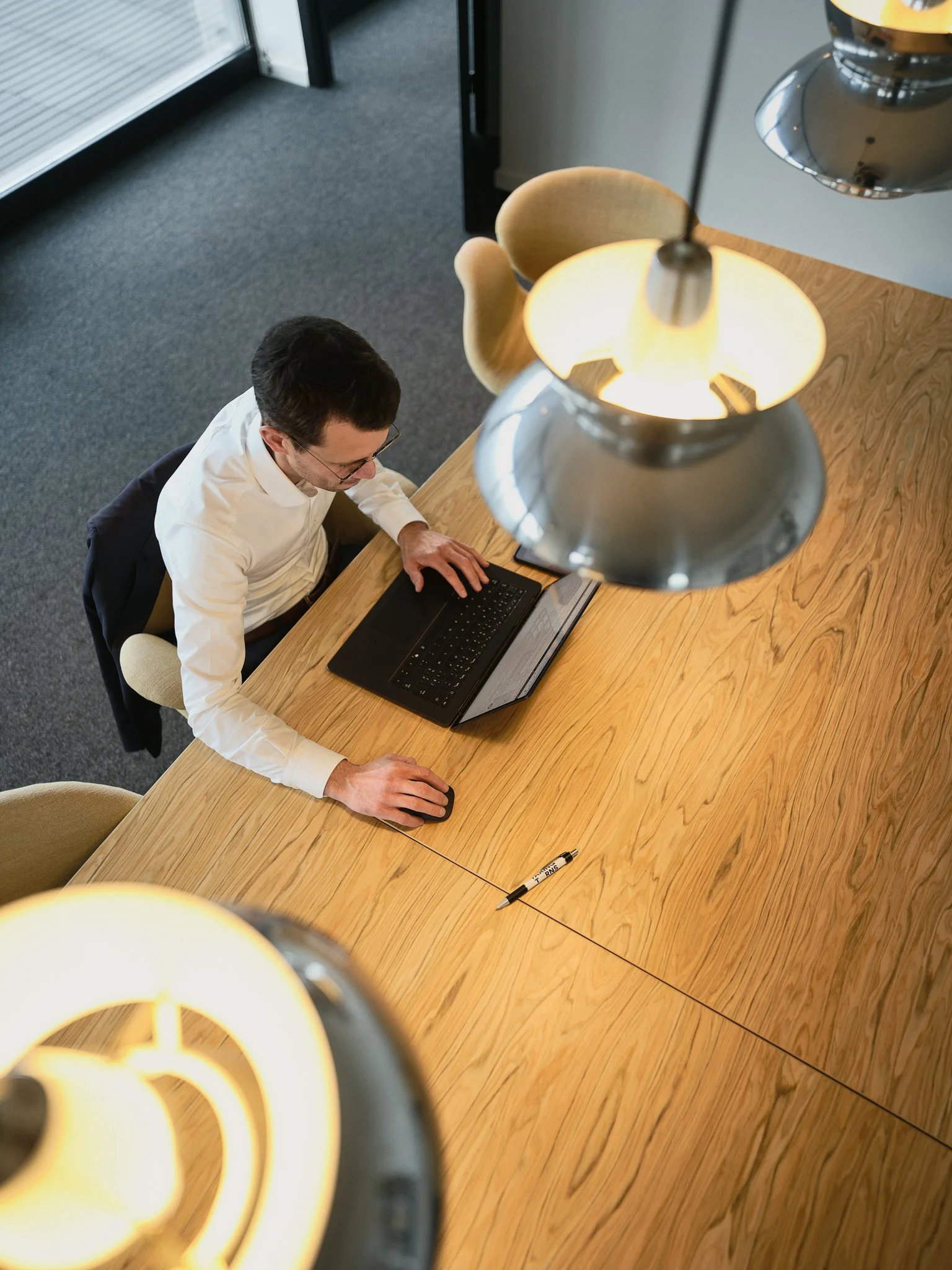 From an overhead view, a man in a white shirt and glasses is working on a laptop at a wooden table, with a black marker nearby. The scene is illuminated by a hanging lamp, and there's a chair and a window with blinds in the background.